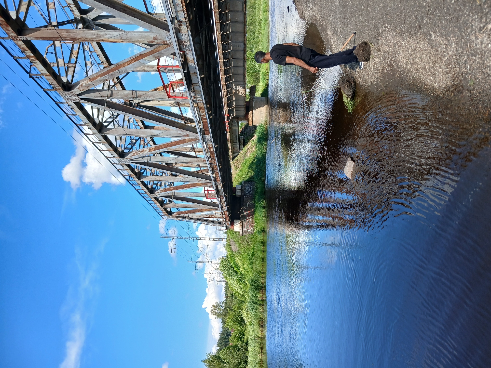 This media file captures a scenic view of a bridge and a river in Yekaterinburg, Russia. The image is oriented vertically, with the sky and the bridge structure occupying the left half, and the river and a person on its bank filling the right half.
The bridge appears to be a railway bridge, constructed from weathered metal beams forming a complex lattice structure. Beneath it, another, older bridge structure is visible, with a more solid, metallic appearance. Electrical power lines are strung across the bridge, suggesting it is an active transportation route. The sky on the left is a vibrant blue, dotted with white, fluffy clouds, indicative of a clear, sunny day. Lush green vegetation, including trees and reeds, lines the riverbank, creating a natural contrast to the industrial elements of the bridge.
On the right side of the image, a person is standing on the concrete bank of a wide, calm river. They are wearing a dark t-shirt and dark pants. The person is partially facing away from the camera, looking towards the water. They appear to be holding a long, thin object, possibly a stick or a fishing rod. The water is a deep blue, reflecting the sky and the bridge structure, with ripples disturbing the surface in places. A small, light-colored rock is visible in the water near the person.
The overall impression is one of a peaceful, natural setting juxtaposed with a significant piece of infrastructure. The bright daylight and clear sky suggest the photo was taken during the daytime. There is no visible text in the image.