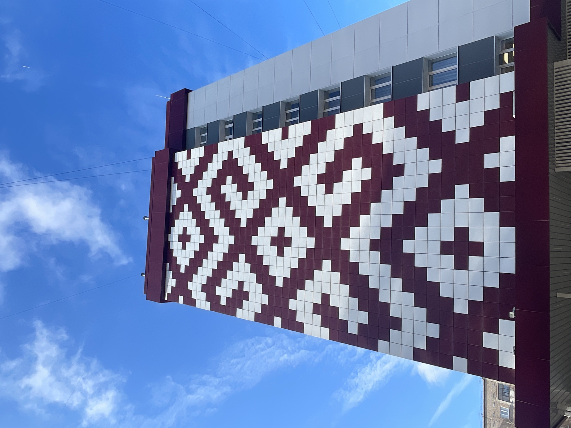The image captures an upward-angled view of a modern building's facade against a clear blue sky with scattered white clouds. The prominent feature is the building's lower section, adorned with a striking geometric pattern composed of alternating maroon (or deep burgundy) and white square tiles. This tessellated design forms interlocking diagonal shapes, reminiscent of traditional folk art or pixel art.
Above this patterned section, the building transitions to a horizontal band of dark grey paneling, featuring evenly spaced rectangular windows. Further up, a lighter grey or white section forms the uppermost visible part of the structure. Thin utility wires crisscross the sky in the foreground, and a faint airplane contrail is visible high in the blue expanse on the left. On the right side of the building, parts of its maroon side paneling and structural elements are visible. The bright sunlight indicates a clear daytime setting. A reflection of another, possibly older building with a window can be faintly seen in the lower right corner, suggesting an urban environment. No people or visible text are present in the image, which is set in Izhevsk, Russia.