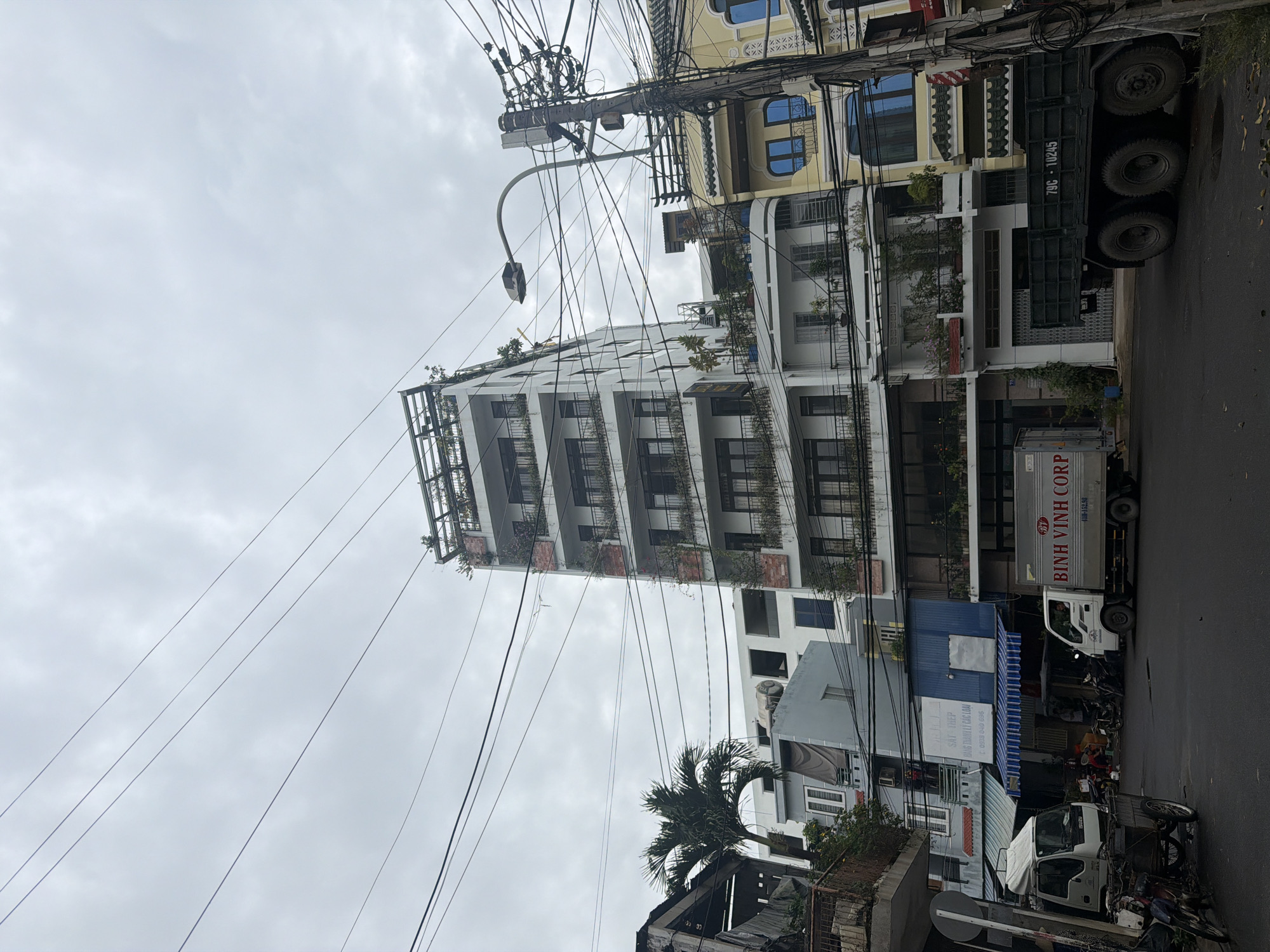 An eye-level street view captures a bustling urban scene in Nha Trang, Vietnam, on an overcast day. The image is dominated by multi-story buildings and a dense web of overhead power lines.
In the center-left, a prominent modern white building rises several stories, featuring multiple balconies adorned with lush green plants. To its right, partially obscured by the power lines, stands a yellow building with distinctive blue-tinted arched windows. Along the street level, a variety of other buildings, including some with blue awnings and signage, are visible.
The street itself, visible in the lower right, has parked vehicles. A large dark green dump truck, bearing the license plate "79C - 10245", is parked on the right side. Further down, a white box truck with "BINH VINH CORP" and "43H - 152.98" printed on its side is also parked. Motorcycles are scattered near the buildings. A street light extends from a utility pole, adding to the complex infrastructure. Palm trees and other greenery provide touches of nature amidst the urban landscape.
The sky is uniformly grey and cloudy, suggesting an overcast day without direct sunlight. The numerous, tangled power lines stretching across the frame are a notable characteristic of the urban setting.
Visible text includes "SẮT THÉP" (Steel), "DẠNG THÀNH LÝ CÁC LOẠI" (Liquidation of all types), and a phone number "0938 040 696" on a blue sign, along with company names and license plate numbers on the trucks.