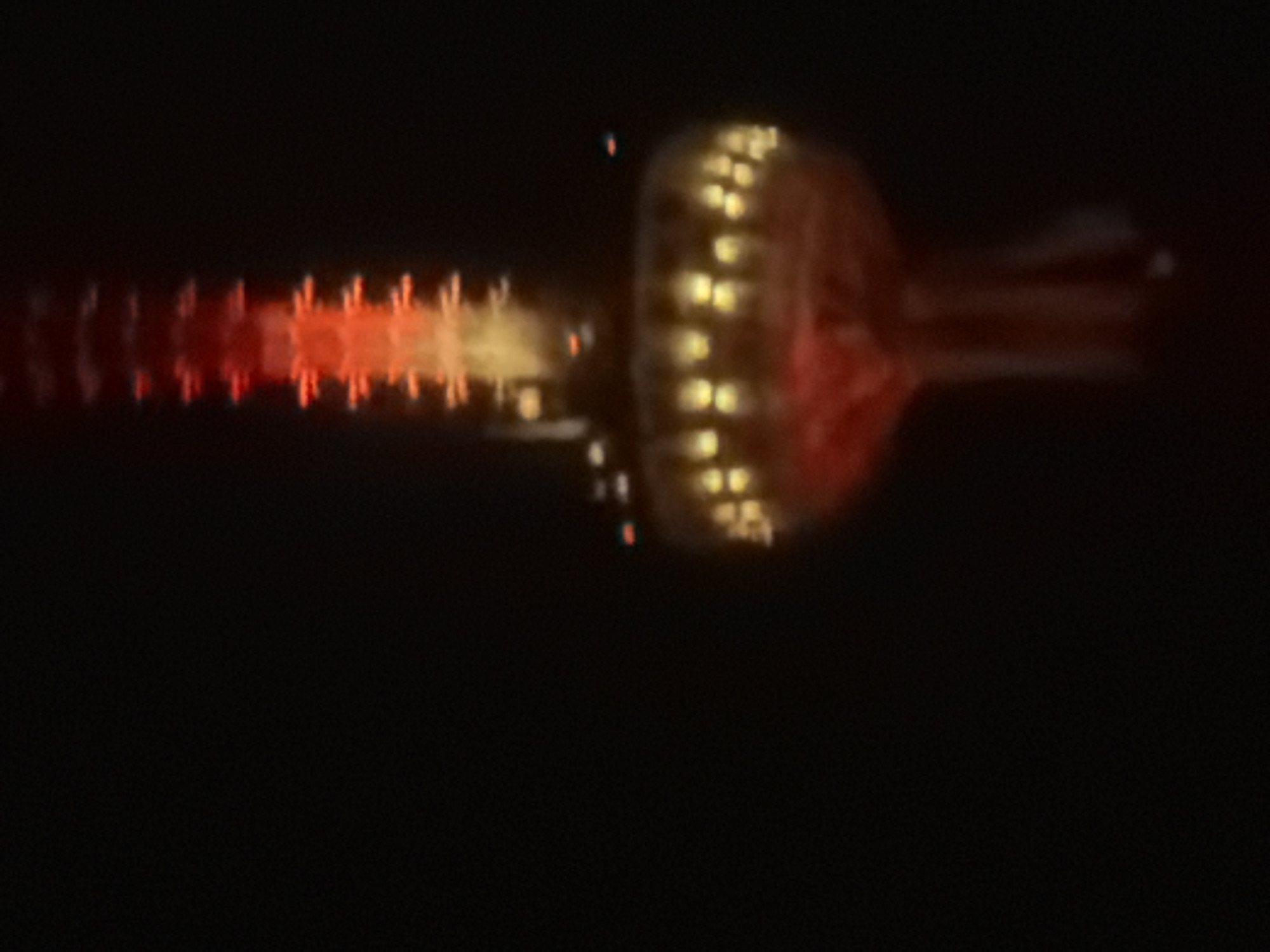 The image shows a blurred, long-exposure photograph of a light display in Tbilisi, Georgia. The display features a central circular component with numerous illuminated dots arranged in a grid-like pattern. Emanating from the left of this structure is a linear series of lights, transitioning from red to orange hues. To the right of the circular section, several light trails extend outward, possibly due to camera movement. The background is predominantly dark, suggesting a nighttime setting.