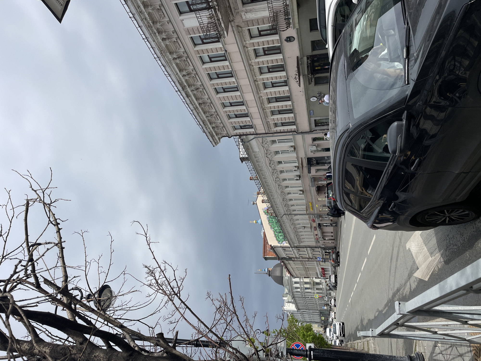 The image shows a street scene in Kazan, Russia, under a cloudy sky. The view is partially obstructed on the right by a black car. The buildings lining the street exhibit classical architectural details. In the foreground on the left, bare branches of a tree are visible. A street sign is present, indicating traffic regulations. In the distance, a building with a dome is visible. The street appears to be relatively empty.