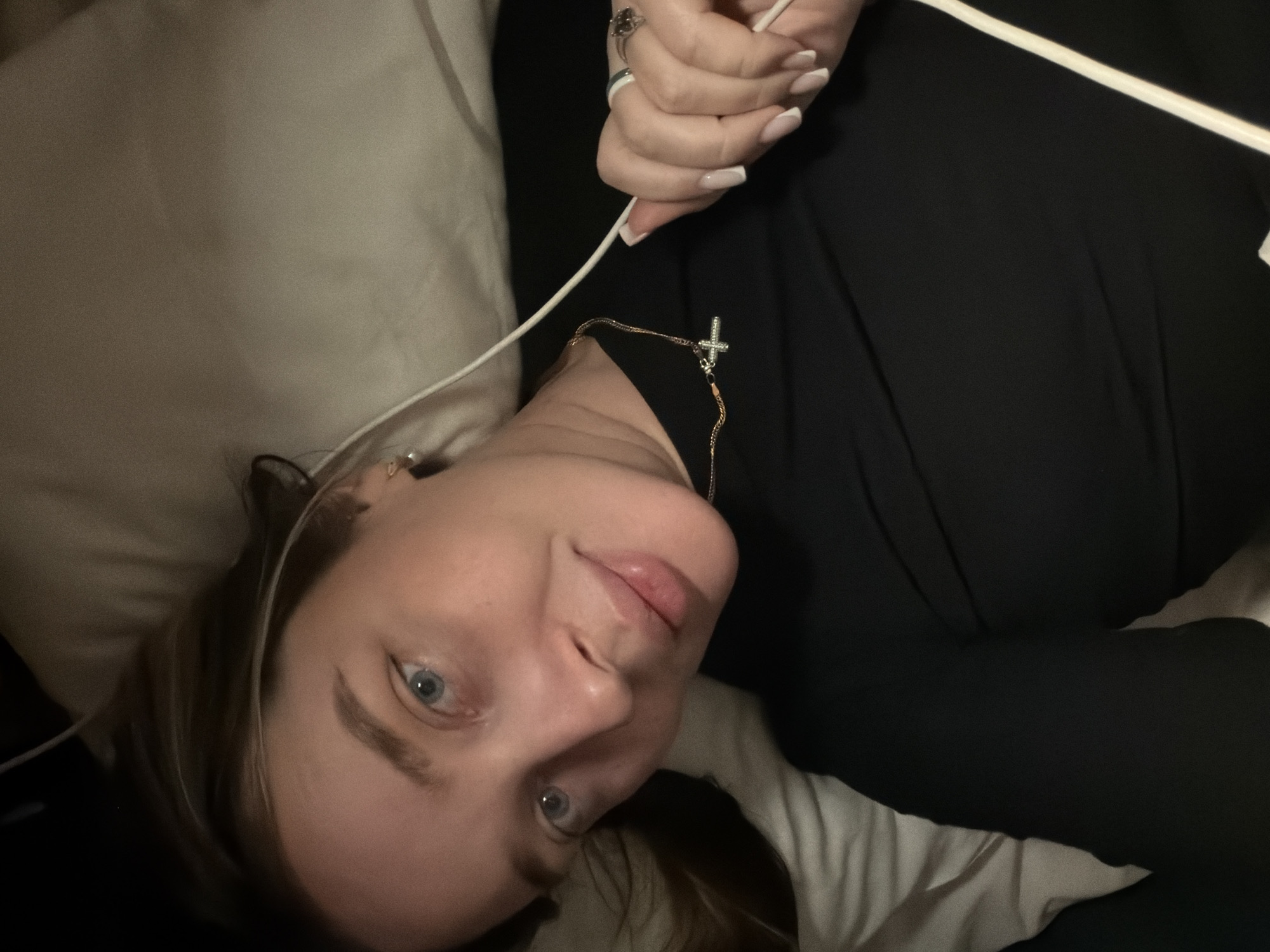 A portrait-style image captures a young woman lying down, angled from her chest upwards, looking directly at the camera with a subtle, relaxed smile. She has light brown hair, fair skin, and engaging blue eyes.
She is wearing a dark, possibly black, long-sleeved top or dress. Around her neck, she wears a delicate gold chain from which hangs a small, silver-colored cross pendant, adorned with what appear to be tiny clear stones. Her left hand is visible in the upper part of the frame, holding a white cable, which could be for headphones or a charger. A ring is visible on one finger of her left hand, and her nails are neatly manicured in a French tip style.
The setting appears to be indoors, likely a bedroom or lounge area, with her head resting on a soft, light-colored pillow or sheet. The background is dark and indistinct, suggesting dim lighting, possibly indicating evening or night. The overall image has a slightly soft or grainy quality. No text is visible in the image. The provided context indicates the location is Moscow, Russia, though no specific visual cues in the image confirm this.