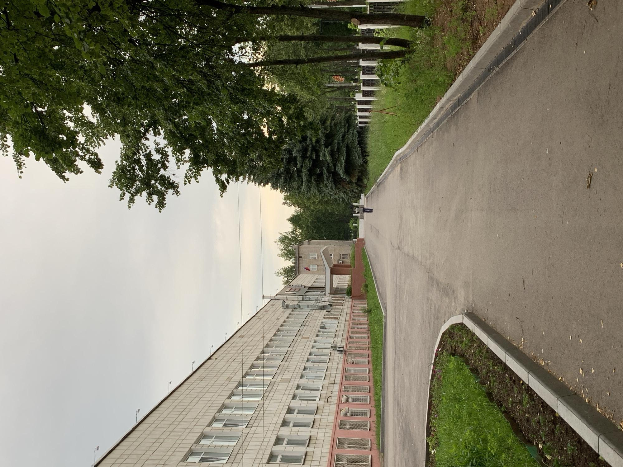 The image shows a view down a paved pathway in front of a long, beige tiled building. The building has many windows and a pink lower section with smaller, barred windows. Green grass and young plants are in garden beds along the side of the building and the pathway. To the right of the pathway, there is a strip of green grass followed by a white fence with dark posts. Beyond the fence are several tall trees, including a large, dark green fir tree and other deciduous trees with lush green foliage. The sky is overcast and grey. A single person is walking away from the camera down the pathway in the distance. The scene appears to be a campus or institutional setting. Based on the architectural style and the presence of a Russian flag on a smaller building in the background, this location is likely in Russia. The image is consistent with the appearance of educational institutions in the city of Chaikovsky, Russia.
