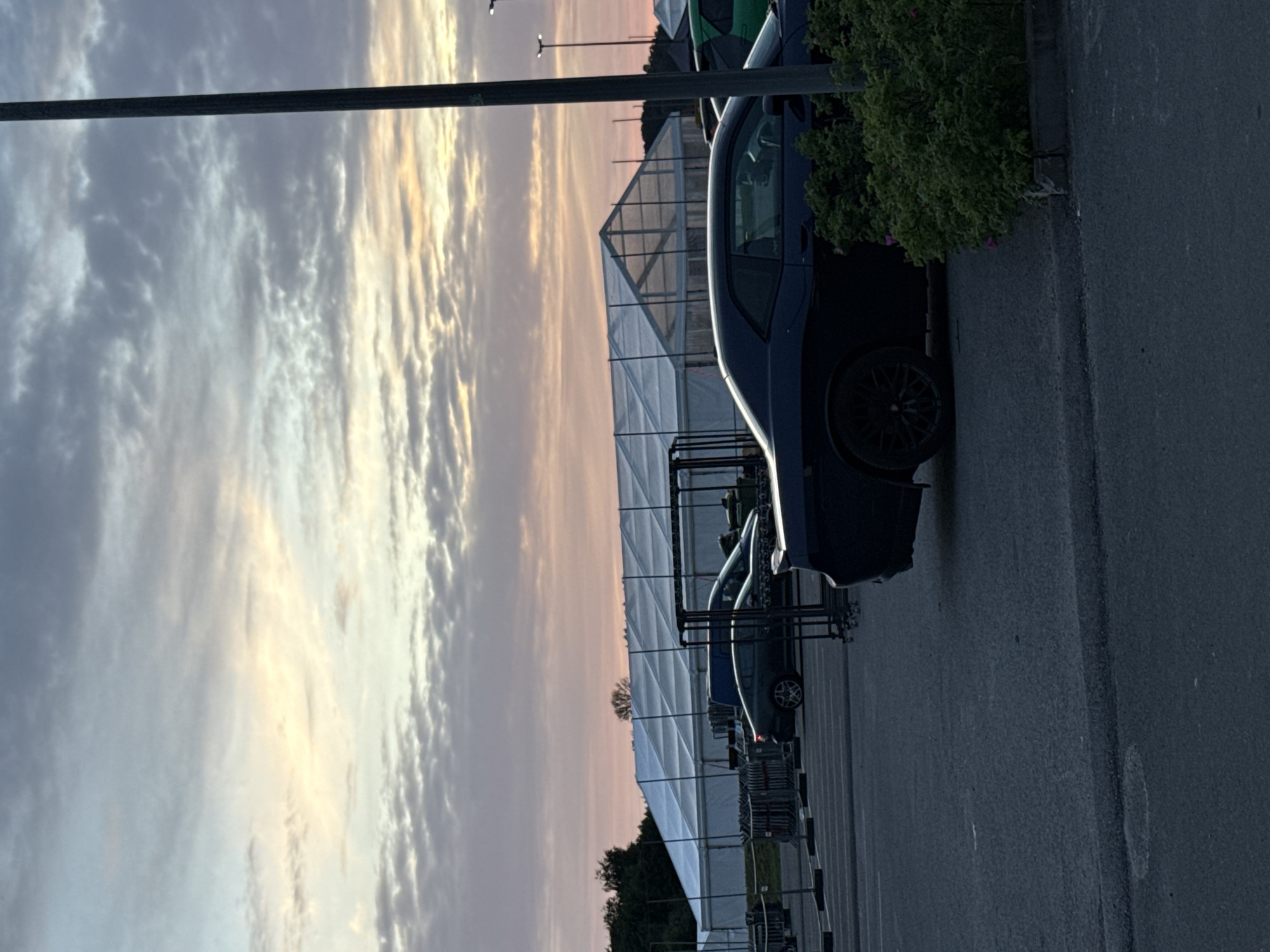 This is a full shot of a parking lot at dusk, with a large white tent structure in the background and a dark blue Dodge Challenger in the foreground. The sky is filled with soft, pastel clouds, indicative of either sunrise or sunset, with hues of pink, orange, and blue. A tall, dark lamppost stands to the right of the frame. In the background, beyond the tent, a treeline is visible, suggesting an outdoor or semi-rural location. The tent appears to be a temporary structure, possibly for an event. Several other cars are visible parked around the tent, and a line of shopping carts is parked to the left. The ground is asphalt. There are no people visible in the image. The scene is calm and still.