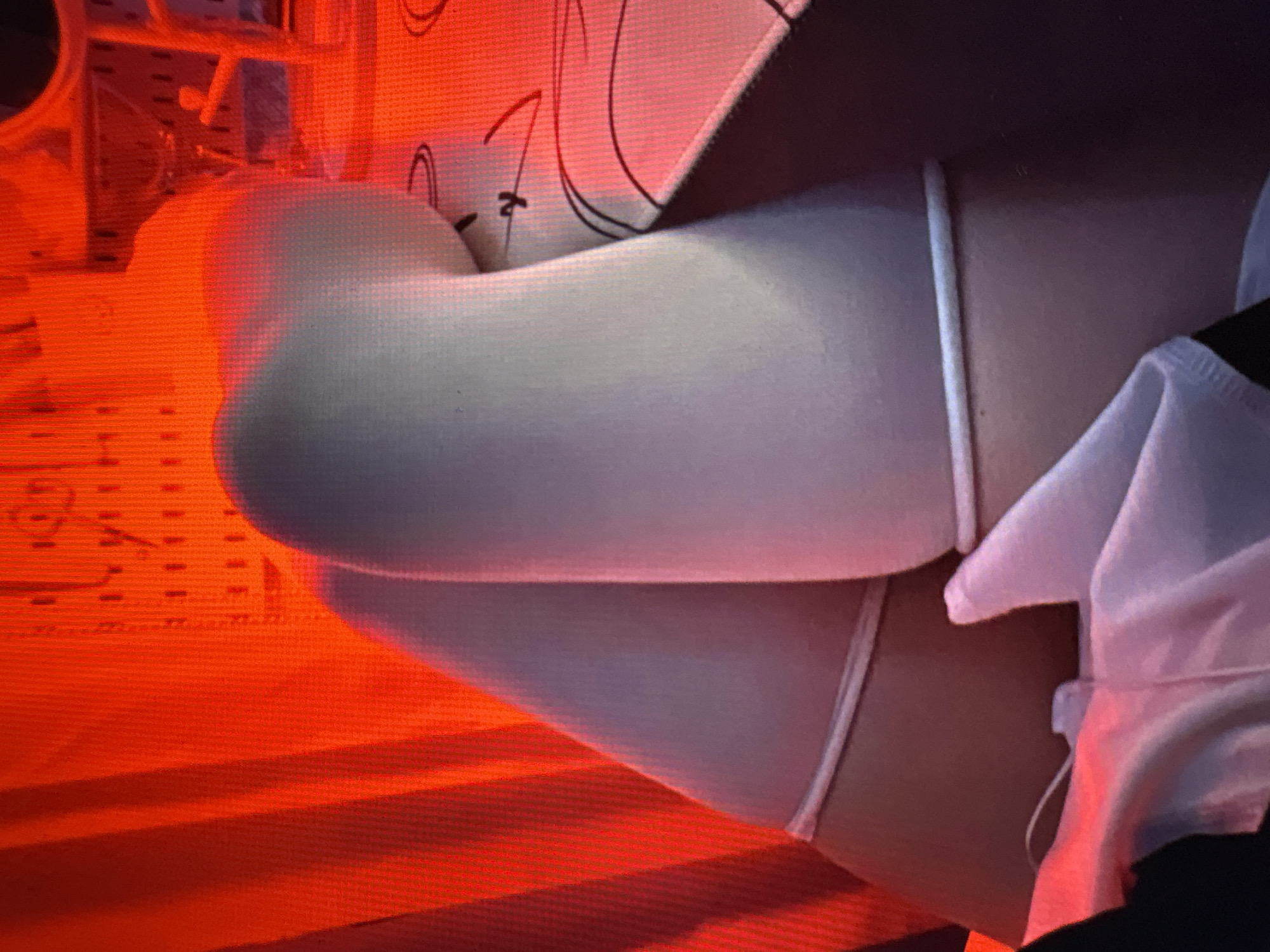 The image shows a close-up view of a person's legs, wearing white thigh-high socks. The socks have a ribbed texture at the top and bottom. The person is sitting on a surface with a patterned design, possibly a table or a bed. The background is dominated by a bright red light, which casts a warm glow over the scene and creates a dramatic, almost surreal atmosphere. The red light appears to be coming from a source behind the person, possibly a lamp or a colored light bulb, and it illuminates the textured fabric of curtains or a backdrop. The lighting is strong and saturated, with some areas appearing overexposed. The overall impression is intimate and artistic, with the focus solely on the legs and the interplay of light and shadow. The specific location within the city of Unknown, Unknown cannot be determined from the image.