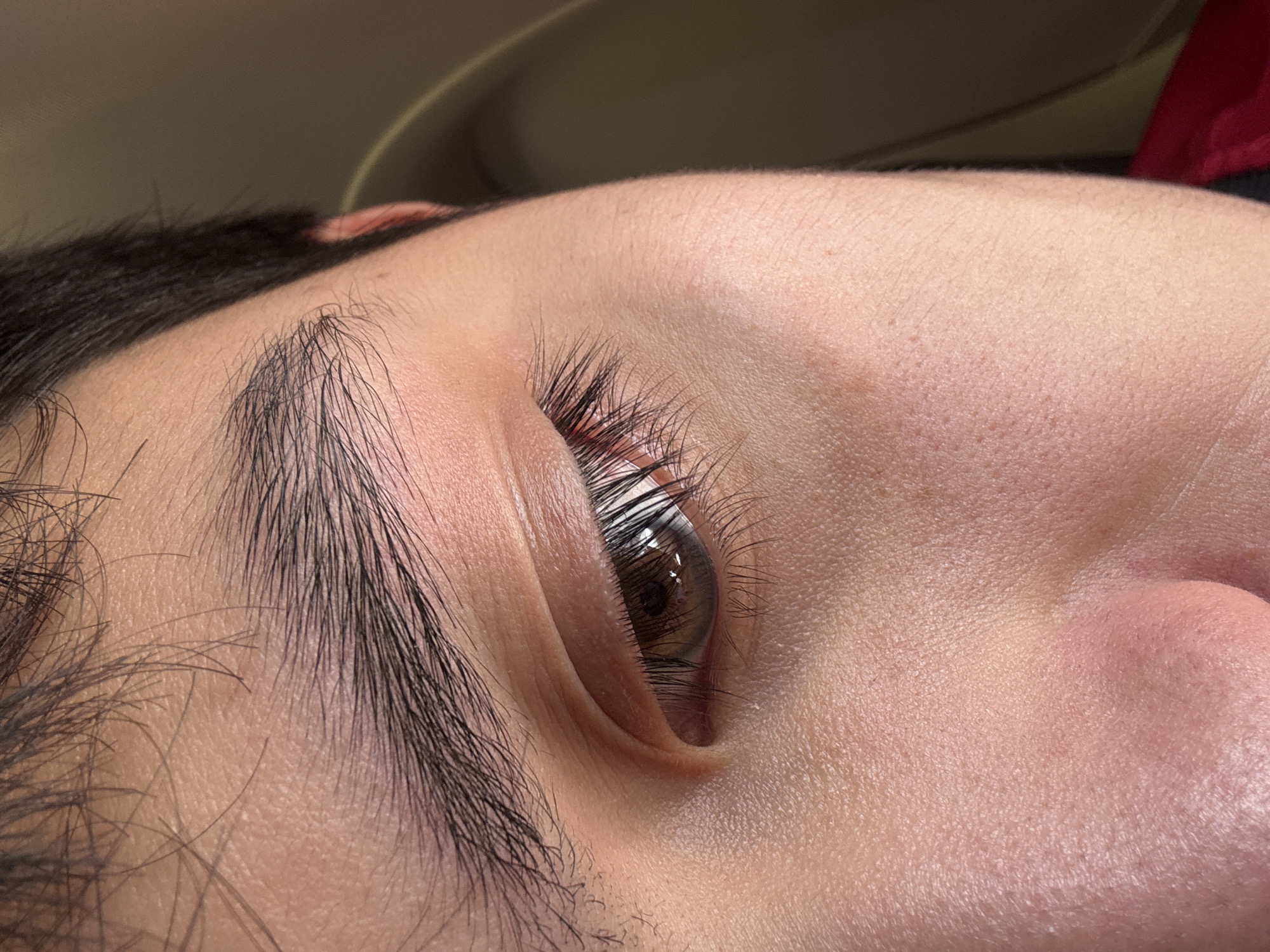 The image shows a close-up view of a person's eye and eyebrow. The eye is light brown with long eyelashes. The eyebrow is dark and full. The skin is fair and appears to have some texture. The background is blurred and out of focus, with a hint of brown and red visible. The image is taken from a low angle, looking up at the eye. There is no discernible information about the location in Mexico City, Mexico.