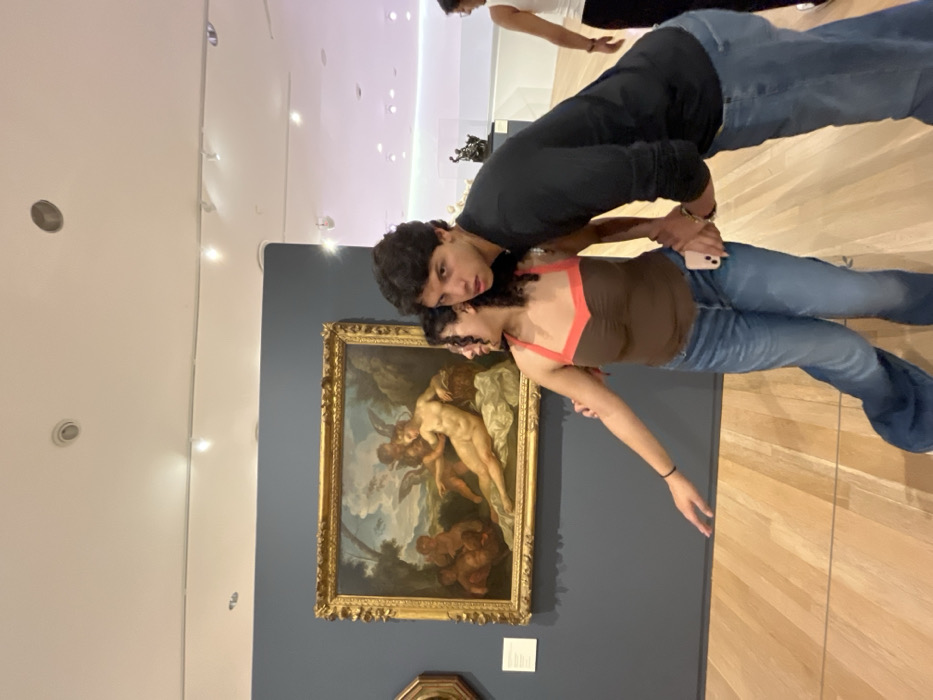 Two people stand in front of a painting. A male wears a black shirt and jeans, looking at the camera. A female, slightly shorter, wears a brown tank top with orange straps and jeans. The painting, in a gold frame, features figures, possibly cherubs and a male nude, in a classical style. The background wall is blue. The floor is wooden, and the lighting is bright. Another person is partly visible in the background.