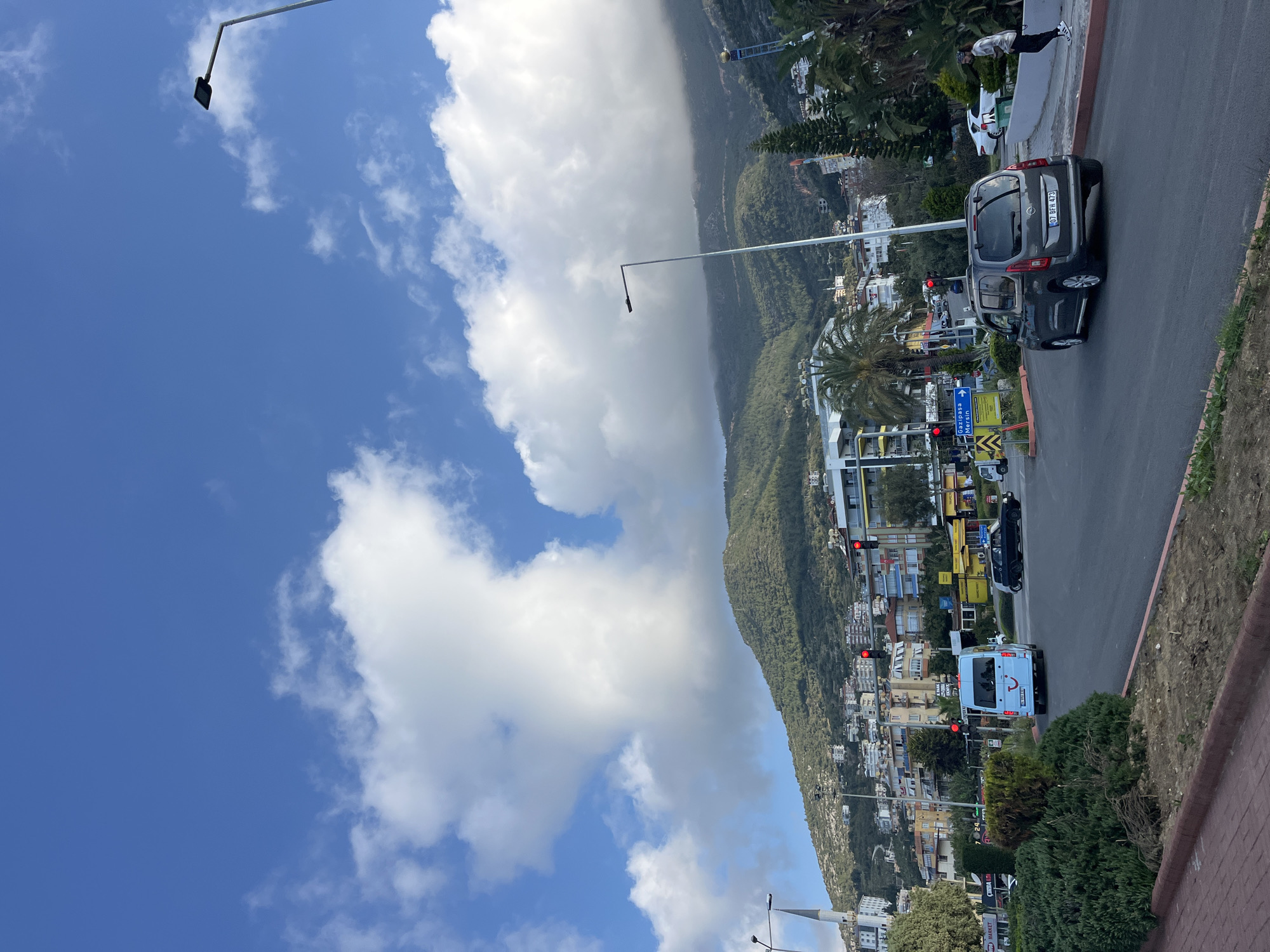 A daytime street scene unfolds in Alanya, Turkey, featuring a busy road flanked by buildings and a large, verdant mountain. The image captures a bright, sunny day under a blue sky adorned with large, fluffy white cumulus clouds.
In the foreground, a multi-lane asphalt road stretches horizontally across the lower third of the frame, gently ascending towards the right. Several vehicles populate the road. Most prominently, a dark grey minivan, possibly a Dacia Dokker, is parked or moving slowly, revealing its Turkish license plate "07 BFH 473". Further down the road, a light blue van, likely a tourist shuttle, is visible from the rear, displaying a distinctive smiling face logo (associated with TUI) on its back door. Red traffic lights are visible at various points along the road, indicating intersections.
The road is bordered by low brick walls, green bushes, and palm trees. Tall, slender lampposts extend into the sky. On the right side, a person in a white shirt and dark trousers stands on a retaining wall next to the road.
Beyond the road, numerous multi-story buildings, typical of a coastal town, line the streets. Some buildings appear to house shops or businesses at ground level. A blue road sign with an arrow pointing left is visible, displaying the text "Gazipaşa" and "Mersin", indicating directions to these nearby cities.
Dominating the background on the right is a massive, tree-covered mountain, its slopes densely forested, creating a striking contrast with the urban development below. The overall impression is that of a vibrant town nestled against a scenic natural backdrop.