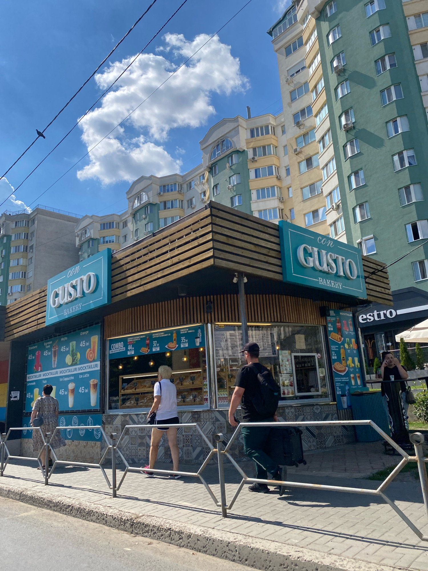 The image depicts a bright, sunny day in Chișinău, Moldova, with a clear blue sky dotted with white clouds. In the foreground, a paved sidewalk runs alongside a street. A metal railing separates the sidewalk from the street. Behind the railing, a bakery named "con GUSTO BAKERY" is prominently displayed with its turquoise and white signage. The bakery has a modern, wooden-slatted facade and large windows showcasing baked goods. A menu board outside displays various drinks and food items with prices in "lei." Several people are visible on the sidewalk. Two women are walking away from the bakery, and a man with a backpack is walking past it, pulling a rolling suitcase. Another person is standing near the bakery, appearing to be on a phone call. In the background, several tall apartment buildings with light-colored facades and green accents rise against the sky, characteristic of urban residential architecture in Chișinău. A pigeon is perched on a power line in the upper left portion of the image.