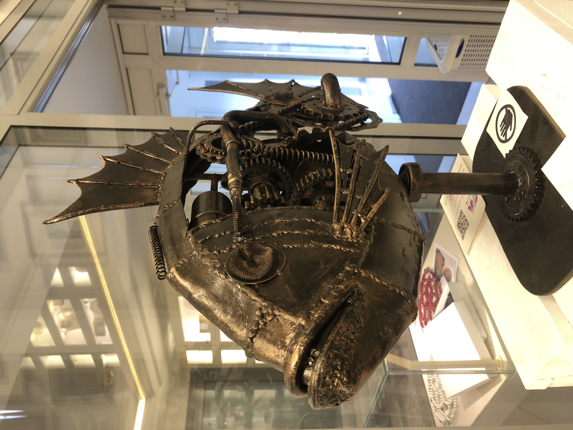 This media file displays a close-up, low-angle shot of a steampunk-inspired metallic sculpture of a fish. The sculpture is predominantly bronze-colored and appears to be constructed from various metal components, including gears, springs, and textured plating, giving it a weathered, industrial aesthetic. The fish sculpture is positioned in what seems to be a display case or shop window, suggested by the reflections and the polished surfaces visible in the background.
The sculpture itself is intricate, with visible gears and mechanical parts incorporated into its design, particularly around the head and body. It has prominent fins that resemble bat wings or mechanical appendages. The fish has an open mouth, revealing the inner workings of its "mechanical" jaw. A coiled spring is attached to its body.
In the background, through what appears to be glass, there are reflections of an indoor environment, possibly a store or gallery. Signs of modern infrastructure, like fluorescent lighting and window frames, are visible. There's also a white triangular display stand to the right of the sculpture, which has a black sticker with a white prohibition symbol (a hand with a diagonal line through it), indicating "no touching." Below this, some paper items are visible, one of which has a QR code and some Cyrillic text. Another item shows a small photograph of a person with a red beaded necklace. The overall lighting suggests an indoor setting, possibly during daytime given the natural light filtering through the windows. The reflections on the glass surfaces and the arrangement of the objects imply a commercial or exhibition space in Izhevsk, Russia.