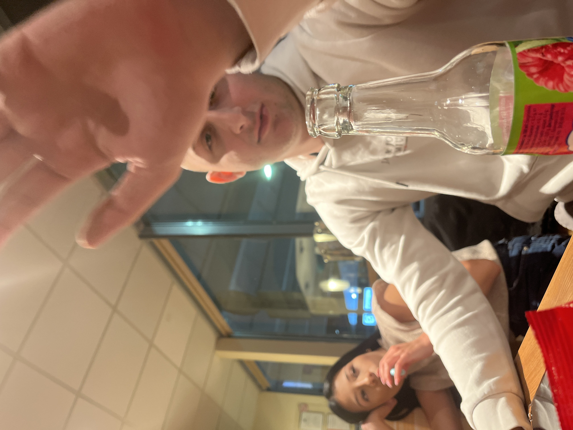 The image shows a close-up perspective of a person holding a glass bottle, with a woman sitting across from them at a table. Both individuals are indoors. The background appears to be a restaurant setting, with windows showing a street scene. Due to the lack of detail of the street scene, it's not possible to establish the location within Burgas, Bulgaria.