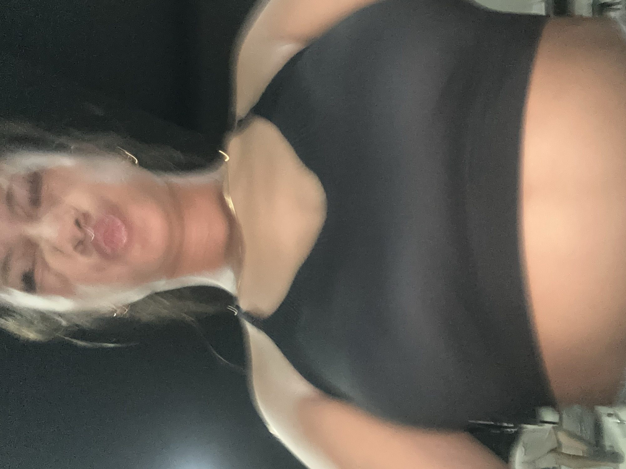 A blurry image of a person, possibly female, wearing a black top and gold jewelry (earrings and necklace). The background is dark, but there are some blurry shapes that suggest the presence of some form of buildings. The image lacks the clarity needed to determine if it was taken in Batubulan, Indonesia, as there are no identifiable landmarks or features to identify its location.