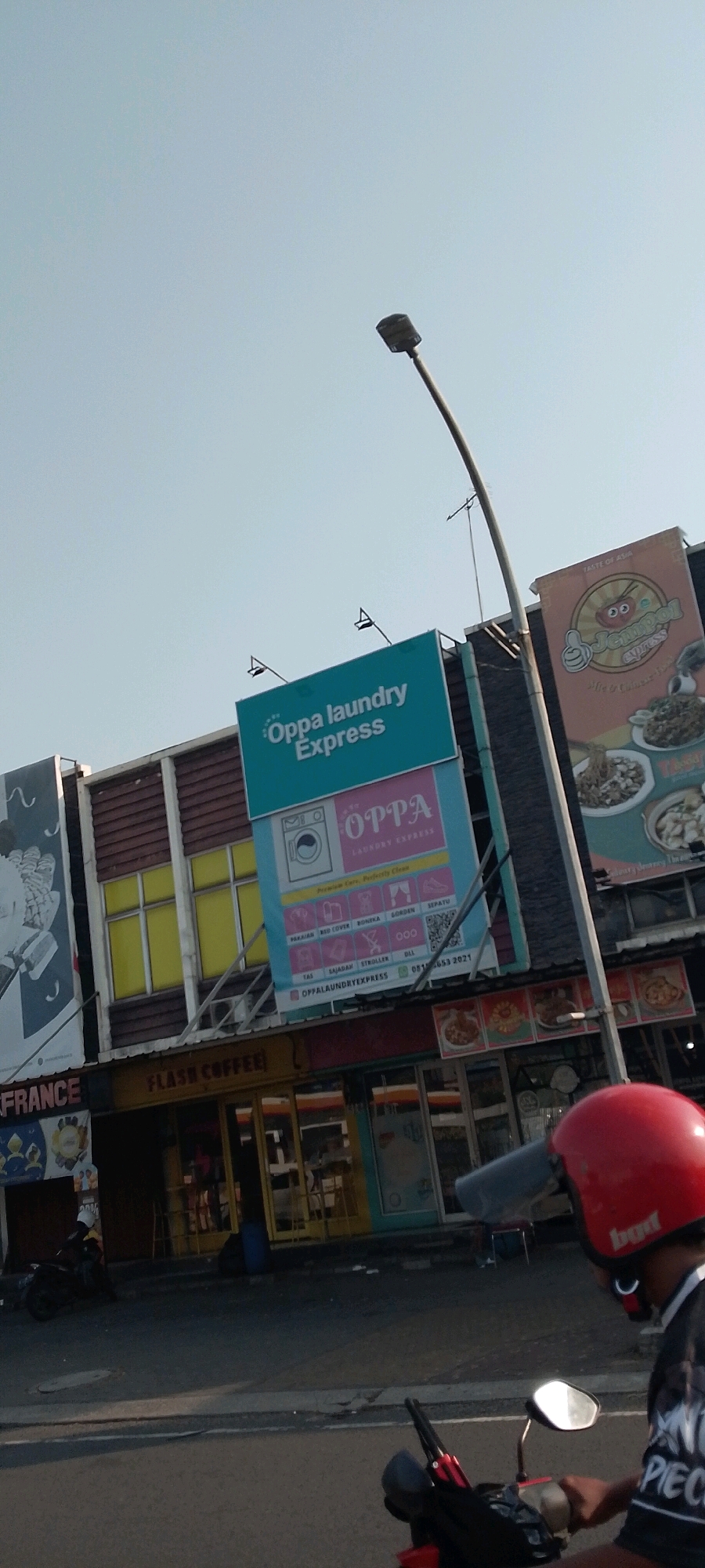 A street-level view captures a bright, sunlit day in Cileungsi, Indonesia. A billboard for "Oppa laundry Express" is prominently displayed on the side of a building. Below it, the storefront for "Flash Coffee" is visible, with its entrance featuring yellow-tinted windows and a dark awning. The street in front of the shops is paved and appears to be an active thoroughfare, with a black motorcycle in the foreground. A person wearing a red helmet is riding the motorcycle, and a second person, partially obscured, is also on a motorcycle. A lamppost extends into the sky from the right side of the frame. The sky is a clear, pale blue.