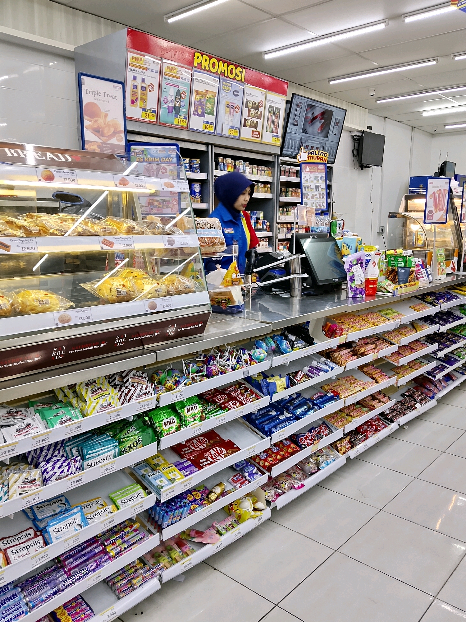 The image captures the interior of a brightly lit convenience store, likely located in Malang, Indonesia, given the Indonesian signage. The foreground is dominated by a long aisle filled with shelves of snack items, predominantly candy bars and packaged sweets, neatly organized and priced. Prominently displayed on the shelves are brands like KitKat, Milo, and Mentos. To the left, a refrigerated display case showcases various baked goods, including buns and pies, also with individual price tags. Behind the counter, a female employee wearing a blue and red uniform, with a headscarf, is visible. She appears to be attending to the register. Overhead, promotional banners advertising deals are displayed. The overall scene depicts a typical retail environment focused on convenience and impulse purchases, with a clear emphasis on food items.