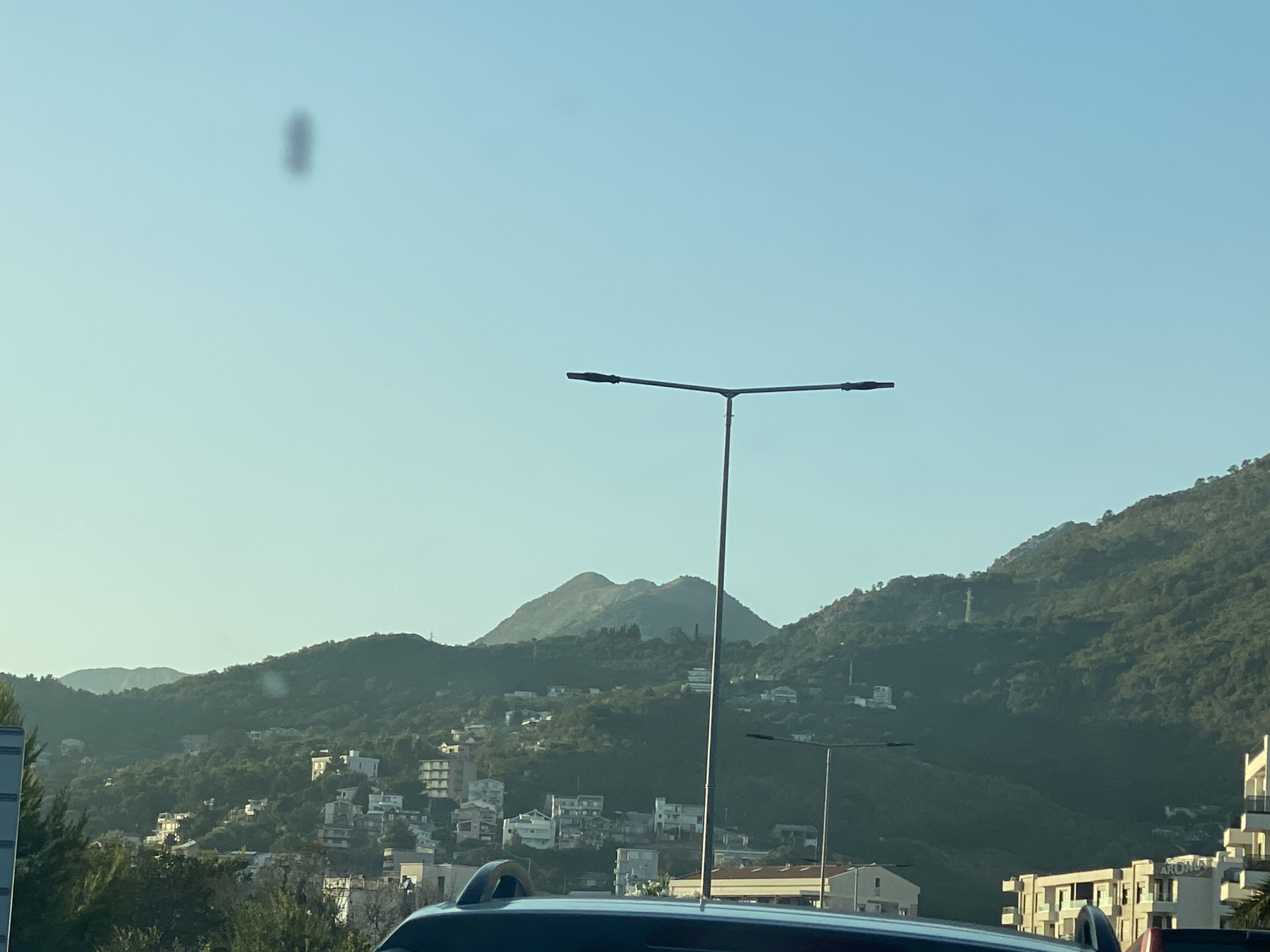 The image captures a view of the city of Bar, Montenegro, with a focus on the mountainous landscape behind it. In the foreground, the roof of a vehicle is visible, partially obscuring the lower portion of the scene. Two modern streetlights with dual lamps stand prominently in the mid-ground, silhouetted against the clear blue sky. The mid-ground also shows a dense scattering of white buildings, characteristic of a Mediterranean settlement, clinging to the lush green hillsides. The hills themselves are covered in dense vegetation, with a prominent, somewhat rounded mountain dominating the background. The lighting suggests it is either early morning or late afternoon, with soft sunlight illuminating the scene and casting subtle shadows. There are no people visible in the frame. The overall impression is one of a coastal city nestled amongst natural beauty.