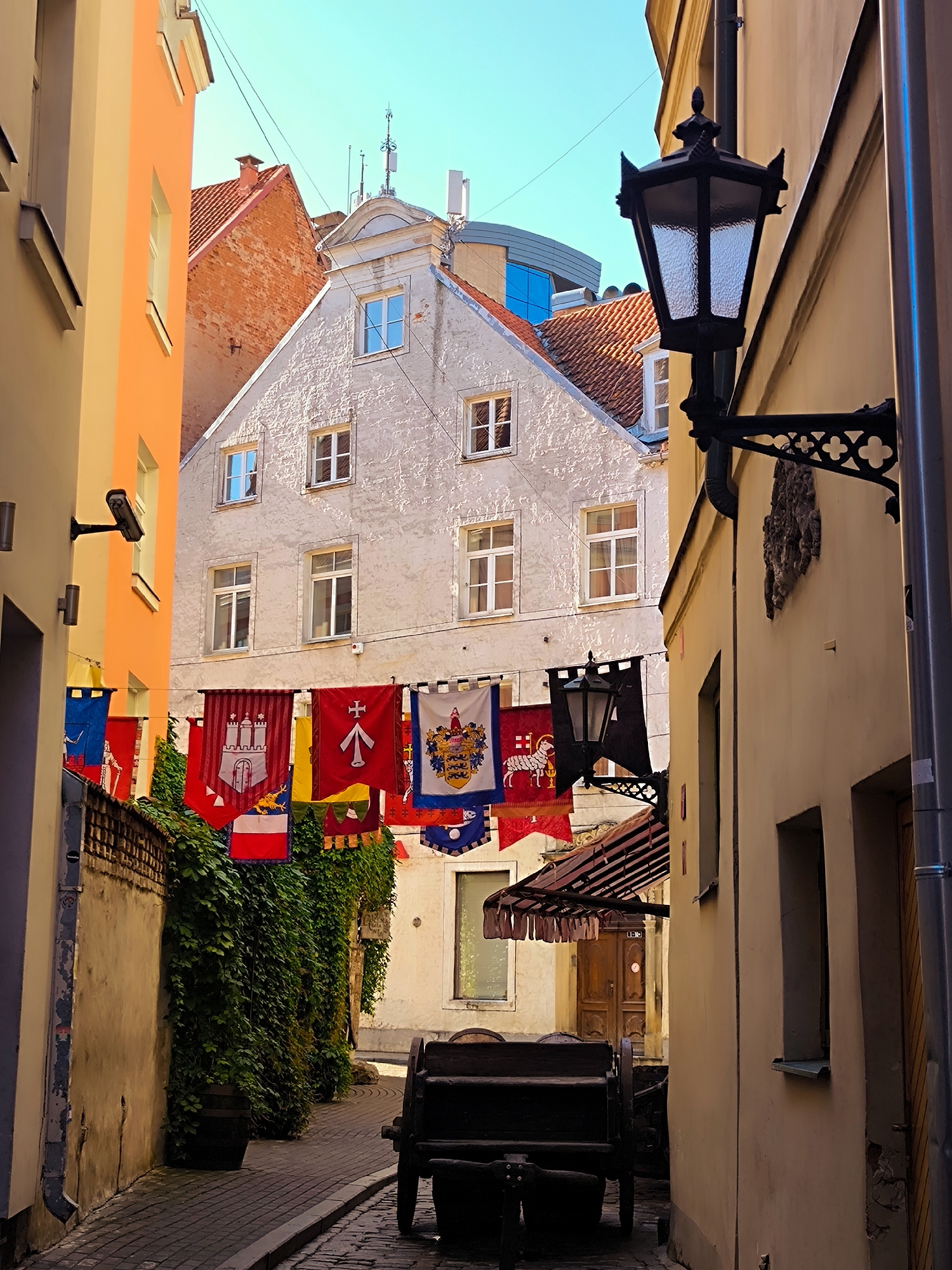 The image depicts a narrow cobblestone alleyway in Riga, Latvia, flanked by historic buildings. The buildings have colorful facades, with one on the left being a pale orange and another in the center a weathered white with red-tiled roofs. Ornate black lampposts are mounted on the walls. A line of colorful banners, featuring various coats of arms and heraldic symbols, is strung across the alley. Lush green ivy covers a portion of the wall on the left. In the foreground, a dark wooden cart with large wheels is parked on the cobblestone street. The sky above is a clear blue. The overall scene suggests an old European city with a medieval or historical ambiance.