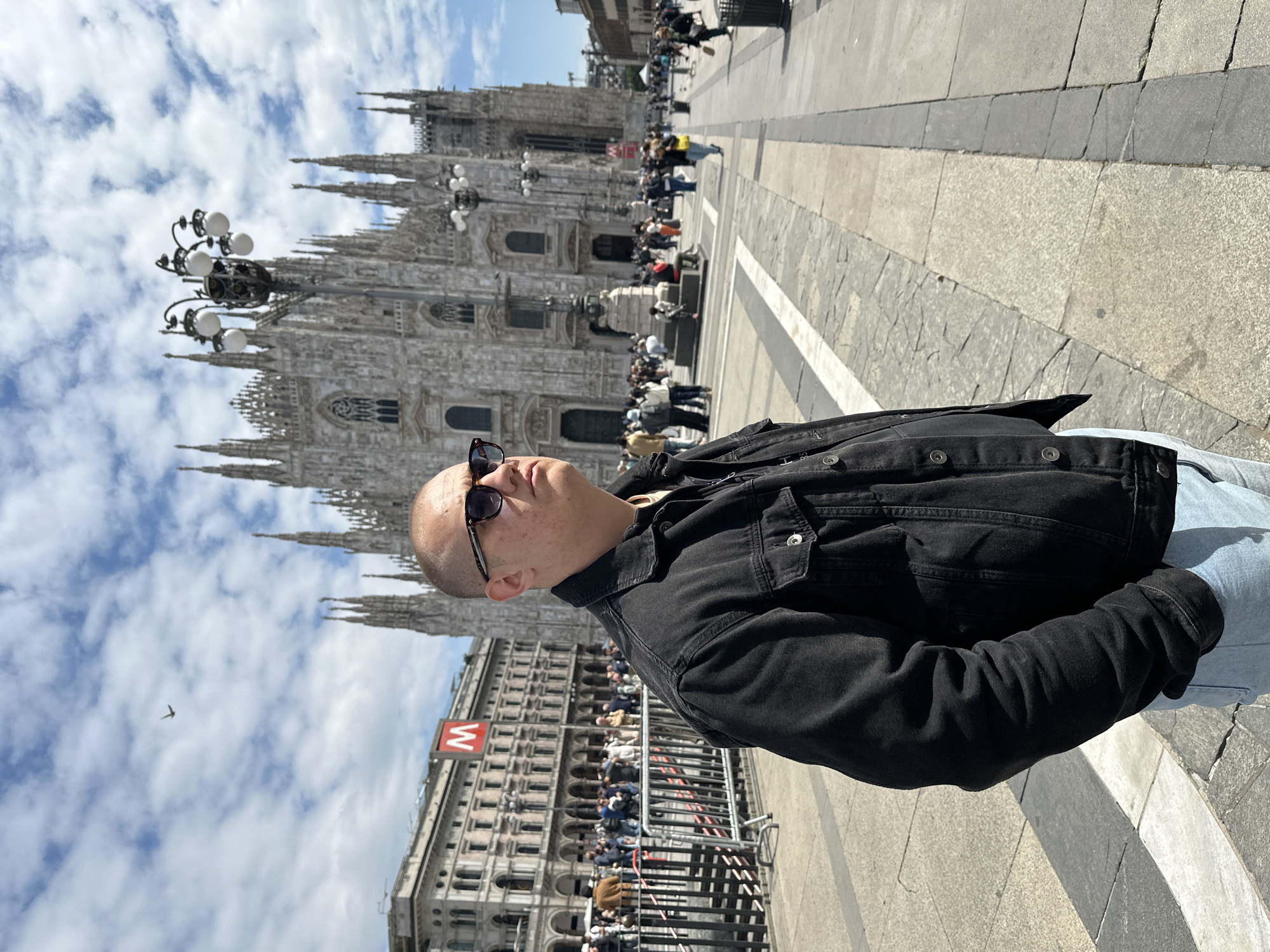 This photograph depicts a sunlit urban scene in a large European city square, oriented as if viewed from ground level. In the foreground, on the right side of the frame, stands a person with a shaved head, wearing dark sunglasses, a black denim jacket, and light-colored jeans. They are looking slightly upward and to the left.
The midground and background feature the impressive Duomo di Milano, a grand Gothic cathedral with numerous spires and detailed architectural elements. To the left of the Duomo, a multi-story building with classical architecture and arched windows is visible. On this building, a distinctive red "M" logo, commonly associated with the Milan Metro, is clearly displayed. A tall, ornate lamppost with multiple globes stands to the left of the main subject.
The square is bustling with activity, as numerous people are scattered throughout, some walking and others congregating near the cathedral. The ground is paved with various shades of grey and white stone tiles, typical of a historic city plaza. The sky is bright with scattered white clouds, indicating a clear, sunny daytime. The strong sunlight casts distinct shadows. The overall setting strongly suggests Milan, Italy, given the iconic Duomo and the metro branding.