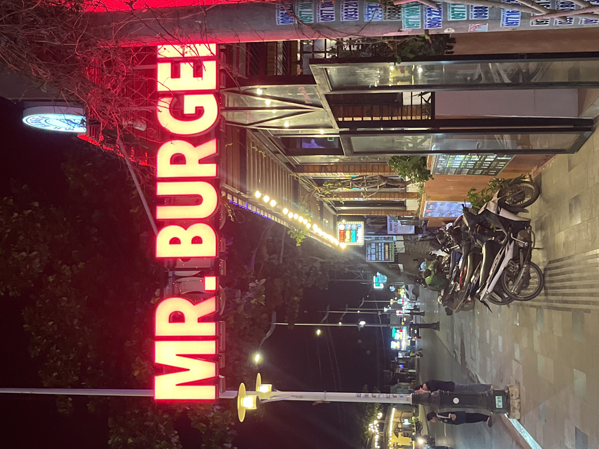 The image depicts a street scene at night in Phu Thuy Ward, Vietnam. The foreground is dominated by a brightly lit neon sign that reads "MR. BURGER." The sign is yellow with a red outline and is mounted above the entrance of a restaurant. To the right of the sign, a circular, illuminated clock with blue numbers is visible.
The street in front of the restaurant is paved with interlocking stones. Several motorcycles are parked along the sidewalk. Two individuals, a person in a dark shirt and jeans and a person in a dark jacket and pants, stand near a lamppost. Further down the street, other vehicles and illuminated signs are visible.
The restaurant itself has large glass windows and appears to be open, with lights illuminating the interior. String lights are draped along the awning of the establishment. The environment is characteristic of a commercial street at night, with artificial lighting from signs and streetlights creating a vibrant atmosphere. The overall impression is that of a lively urban setting.