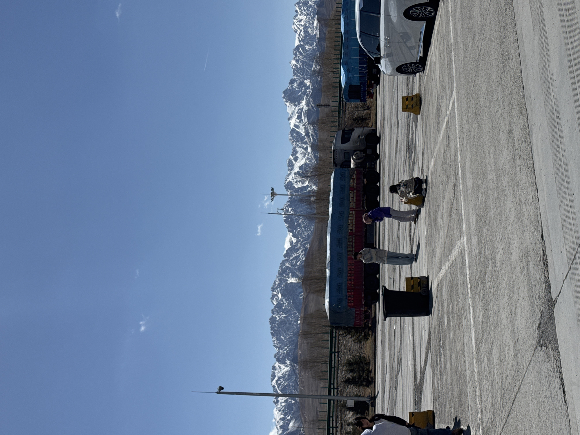 The image captures a parking area in what appears to be a mountainous region, presumably near Wuwei, China. Snow-covered mountains form the backdrop, with a clear blue sky above. A line of parked vehicles, including trucks and cars, is situated on the right side of the frame. Several people are visible in the parking area; some are standing near the trucks. The ground is asphalt, marked with white lines denoting parking spaces. There are yellow traffic cones. A utility pole extends upward on the left side of the image.
