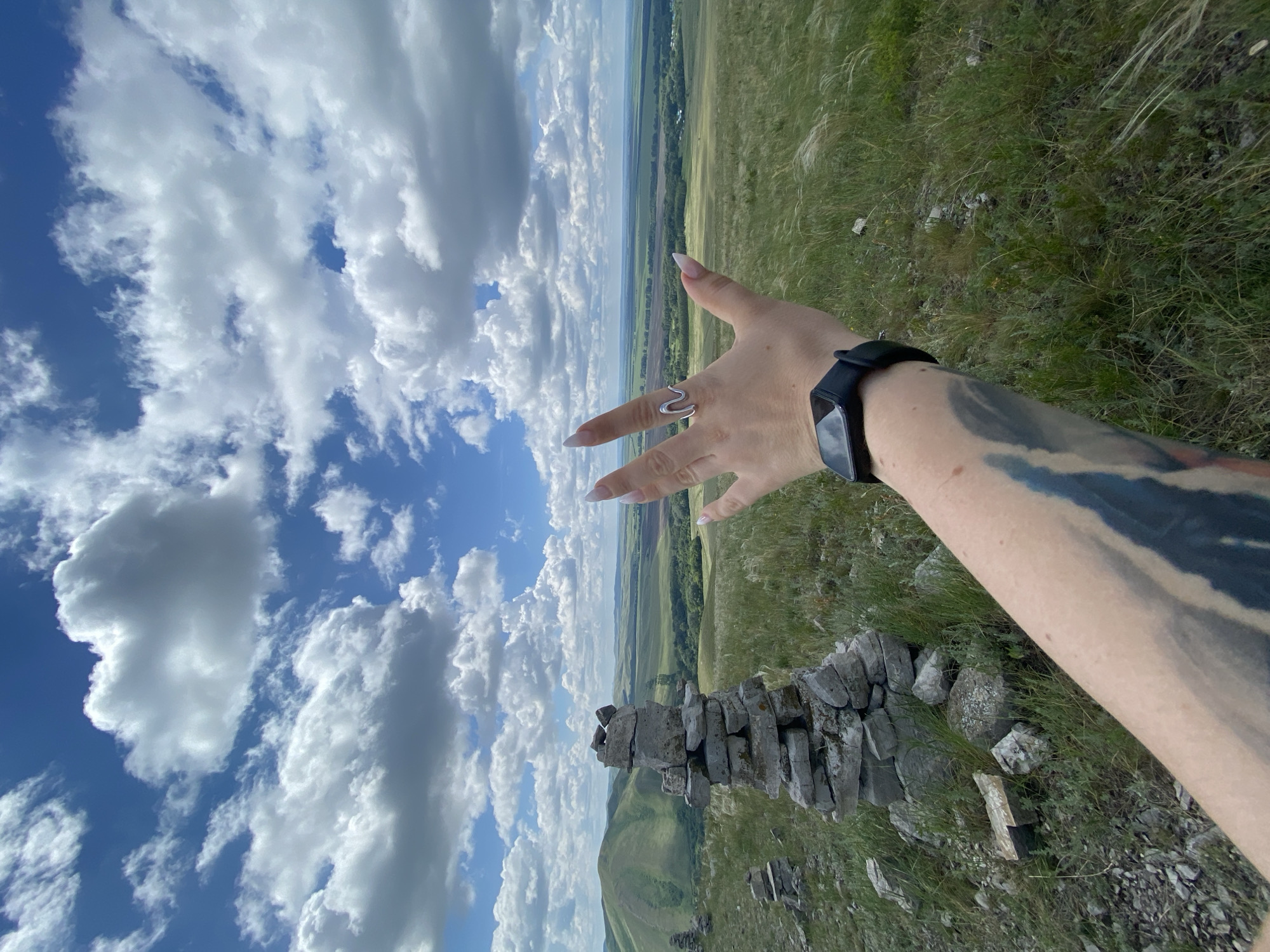 The image captures a scenic view from a hilltop overlooking a vast valley. In the foreground, a person's tattooed arm extends into the frame, wearing a black smartwatch. Their hand, with long, manicured fingernails, is raised and open, adorned with a silver ring shaped like a wave. To the left of the hand, a cairn constructed from stacked stones stands prominently. Behind it, more stone markers are visible. The landscape features rolling green hills, fields, and a line of trees in the distance, all under a bright blue sky dotted with fluffy white clouds. The location is identified as being near Yuldybaevo, Russia.
