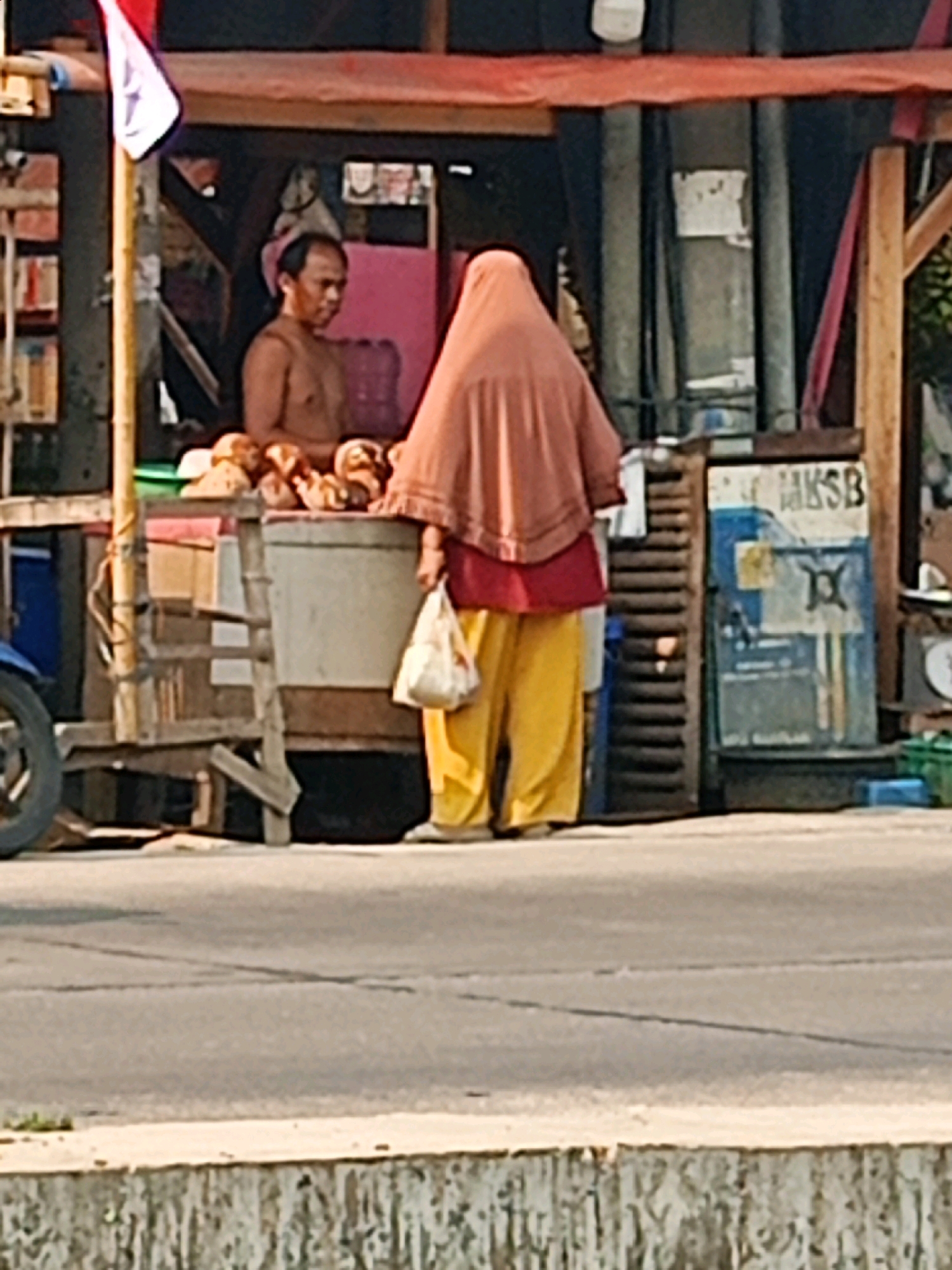 In Sriamur, Indonesia, a scene unfolds at a roadside stall. A man, shirtless and with short, dark hair, stands behind a display of round, bread-like items. He appears to be a vendor. To his right, a woman, wearing a long, rust-colored head covering and bright yellow pants, holds a white plastic bag in her left hand. She is facing away from the camera, towards the bread display. Behind the woman, a stall structure is visible, constructed with wood and metal. A sign with blue and white lettering, including the letters "MKSB," is affixed to a vertical surface. To the left of the stall, the rear wheel of a blue motorcycle is partially visible. The ground in front of the stall is paved with asphalt, with a concrete divider running along the edge. The overall environment suggests a bustling street or market setting.
