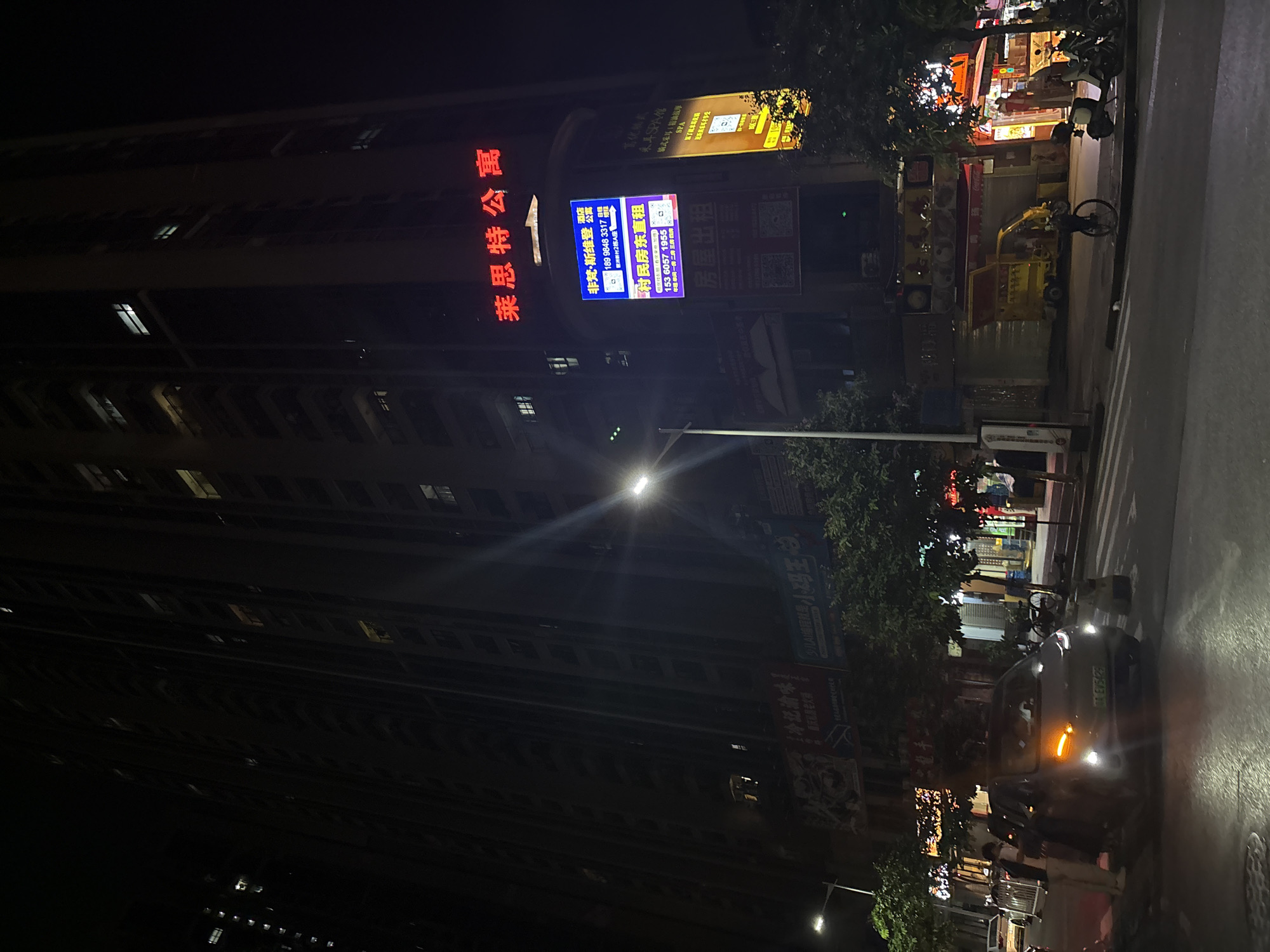 This is a nighttime photo taken in Haizhu District, China. The scene depicts a street with a tall building on the left and shops on the right. The building appears to be an apartment complex, with many windows, some of which are illuminated.
Several signs are visible. A large red sign on the building reads "莱思特公寓" (Laithwaite Apartment). Below this, a blue sign with white and purple text advertises "非梵·斯维登" (Fei Fan · Svidon), a rental service, with a phone number and address. Another sign features "重庆老火" (Chongqing Old Fire), suggesting a restaurant. Further to the right, more signs are present, including one for "東耳SPA館" (Dong Er SPA Museum) offering ear spa services and massages.
The street is paved and appears to be an urban setting. A car with its hazard lights on is parked on the right side of the road, and several motorcycles are also visible parked along the sidewalk. Pedestrians are present near the car. Trees line the street, adding greenery to the urban landscape. The time of day is clearly night, with artificial lighting from the building, shops, and streetlights illuminating the scene. The weather appears to be clear, as there are no indications of rain or fog. The overall impression is a bustling urban environment at night, with commercial activity and residential living coexisting.