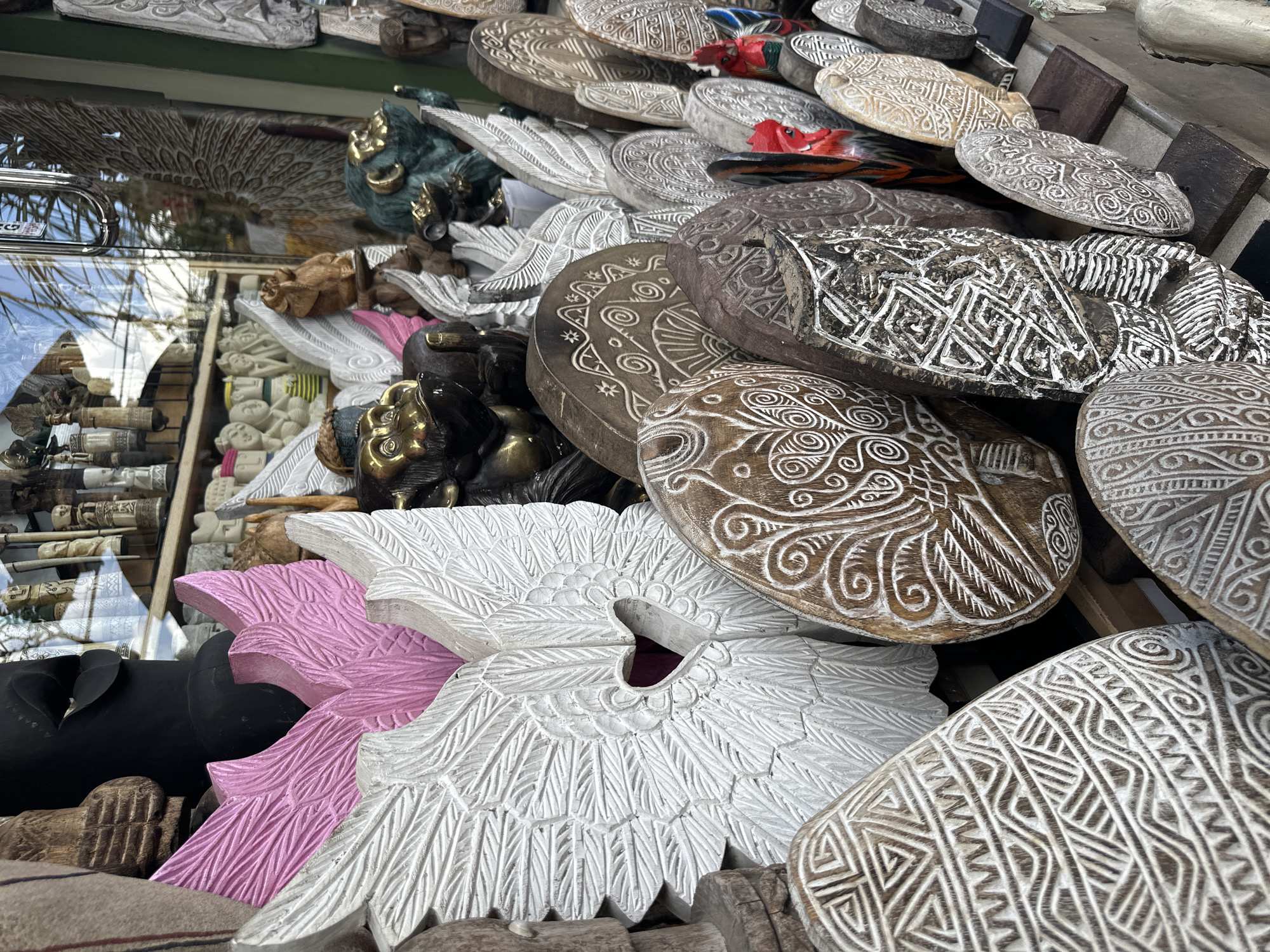 This is a close-up, eye-level shot of a display of various carved wooden crafts, likely from a market stall in the Ubud District, Indonesia. The foreground is dominated by intricately carved circular wooden objects with white geometric patterns and swirls. To the left, a large, white carved wing with a heart-shaped cutout is prominent. Behind it, a pink carved wing shape is visible. Further back, there are more carved wings and several carved animal figures, including what appear to be stylized monkeys with gold accents, and some green animal figures. The background shows reflections of palm trees and the sky in what seems to be a shop window, suggesting the stall is located within or adjacent to a building. The overall impression is one of detailed artistry and a vibrant cultural display of handcrafted goods.