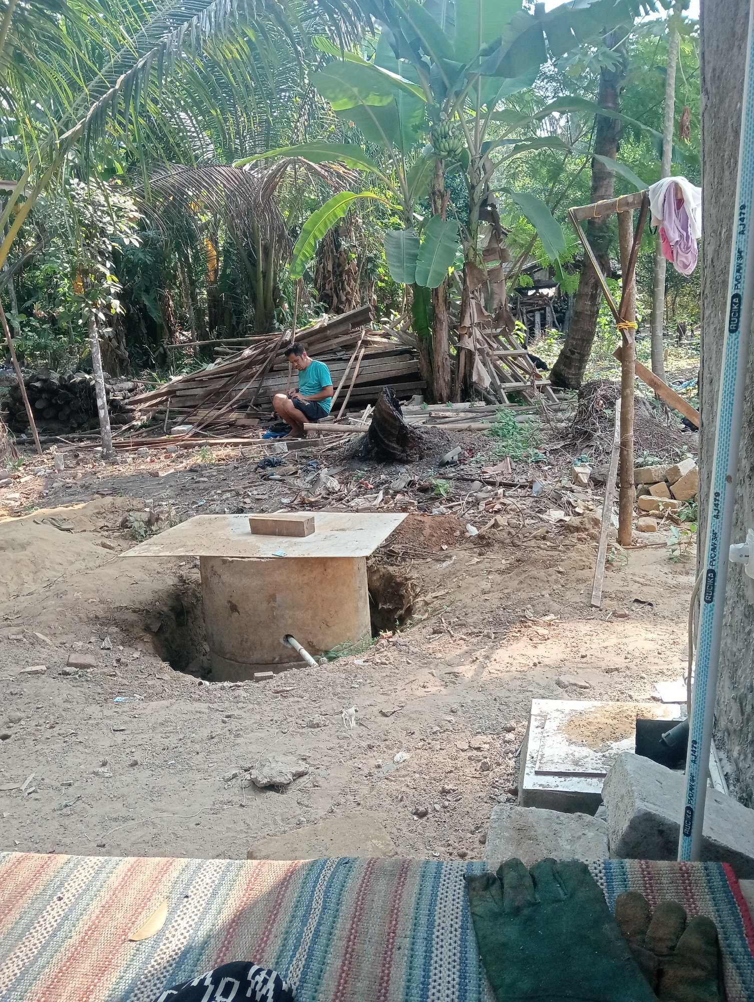 The image depicts an outdoor scene in a tropical, rural environment, likely in Muara, Indonesia, characterized by lush vegetation, including palm and banana trees, and a rustic construction area. In the foreground, a circular concrete structure, possibly a well or septic tank, is partially excavated from the sandy soil. A white pipe protrudes from its side. A man in a turquoise t-shirt and dark shorts is seated on a wooden plank in the midground, looking down at his phone. Behind him, a large pile of lumber is stacked against a backdrop of dense foliage and trees. A makeshift clothesline with two items of clothing hangs from a wooden frame to the right of the man. The ground is covered in dirt, sand, and scattered debris, suggesting ongoing construction or renovation. A close-up of a woven mat with green work gloves is visible in the bottom right corner. The overall impression is one of a utilitarian, rural setting undergoing some form of development.