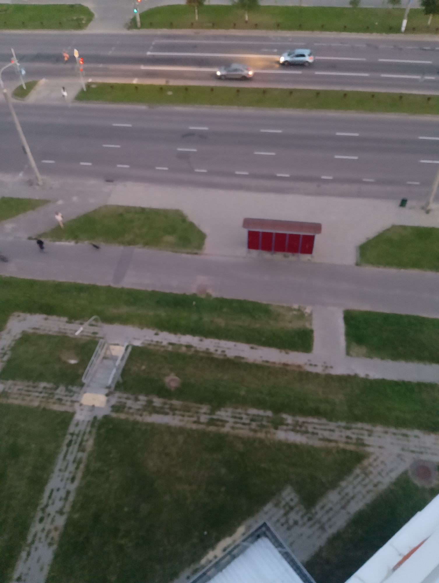 The image shows a street scene in Vitsebsk, Belarus, likely taken from an elevated vantage point such as a building. In the foreground, a grassy area with paved pathways and some greenery is visible. A metal staircase leading to a paved area is also present. Further back, a road with multiple lanes and traffic markings runs horizontally across the frame. Several vehicles are on the road, including two cars moving from left to right. On the side of the road, there is a bus stop shelter with a red roof and side panels. People are walking on the sidewalk and grassy areas adjacent to the road. The overall lighting suggests it is either early morning or late evening.