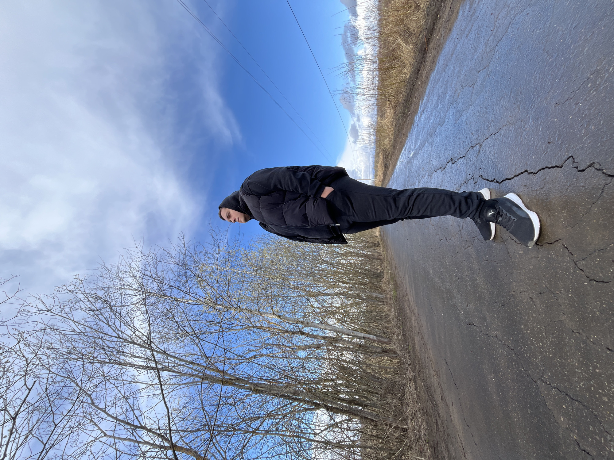 A full-body shot depicts an individual standing on a cracked asphalt surface in what appears to be an outdoor environment. The individual, dressed in black attire, including a jacket and pants, has their hand in their pocket. The asphalt surface has visible cracks and appears slightly damp. To the right of the asphalt, there is a strip of vegetation. On the left side, bare trees reach towards the sky. Above the individual, there is a blue sky with scattered white clouds. Overhead power lines stretch across the upper portion of the frame.
The location is Opalevo, Russia.