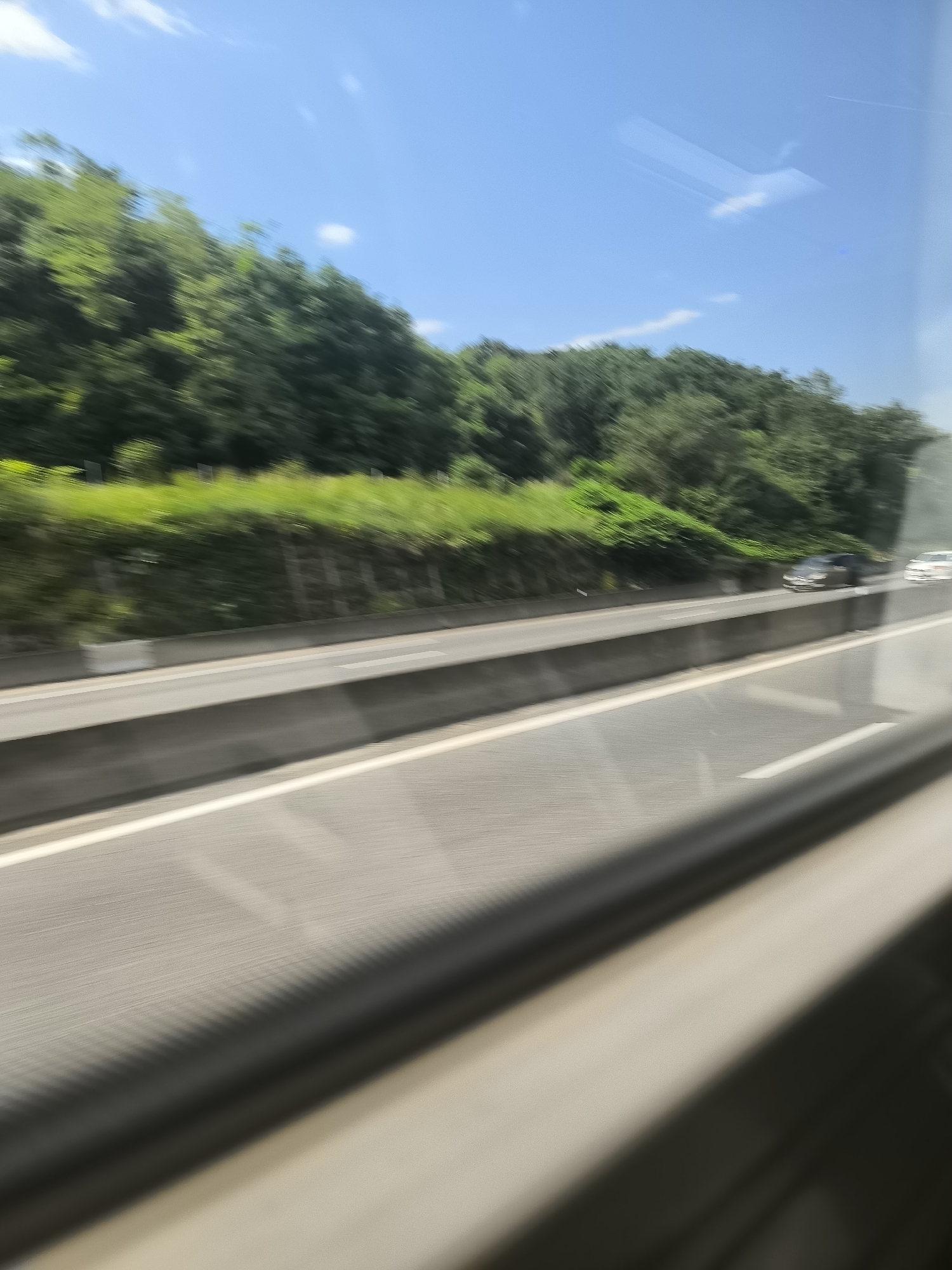 A blurry, moving shot from inside a vehicle on a highway. The view includes a green, tree-covered hillside alongside the road. There are multiple lanes on the highway and cars are driving in the distance. The sky is bright blue with a few clouds. The foreground shows the interior of the vehicle. The location is Saint-Chamond, France.