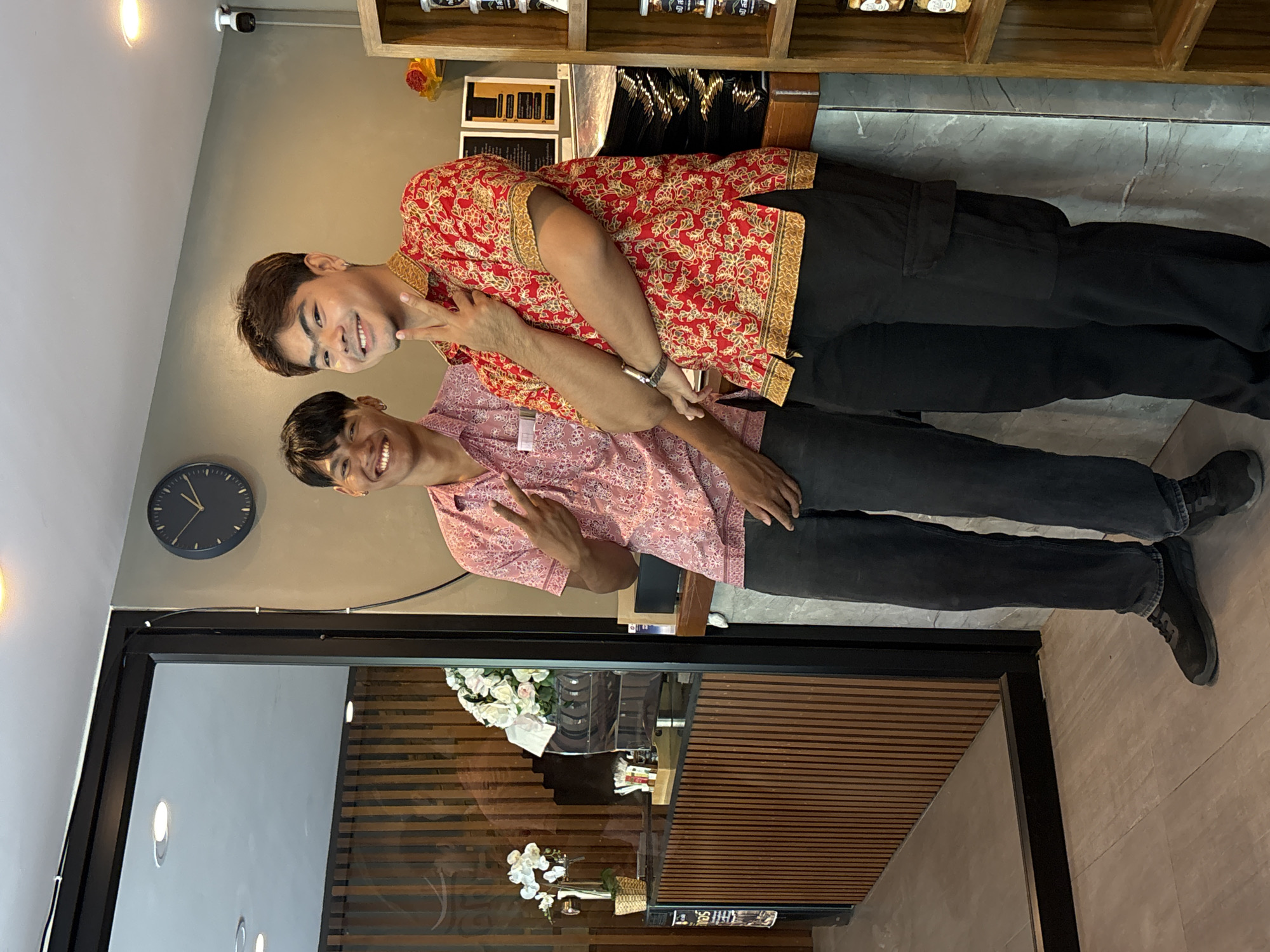 Two men are posing indoors, making peace signs with their hands. The man on top wears a red and gold patterned shirt and black pants, while the man below wears a pink patterned shirt and black pants. Behind them is a wall with a clock. To the left, a doorway reveals a counter or table with horizontal wood slats and white flowers. The scene is in บ้านบางรักษ์, Thailand. Behind the men is a shelf with various items. The floor is light colored.