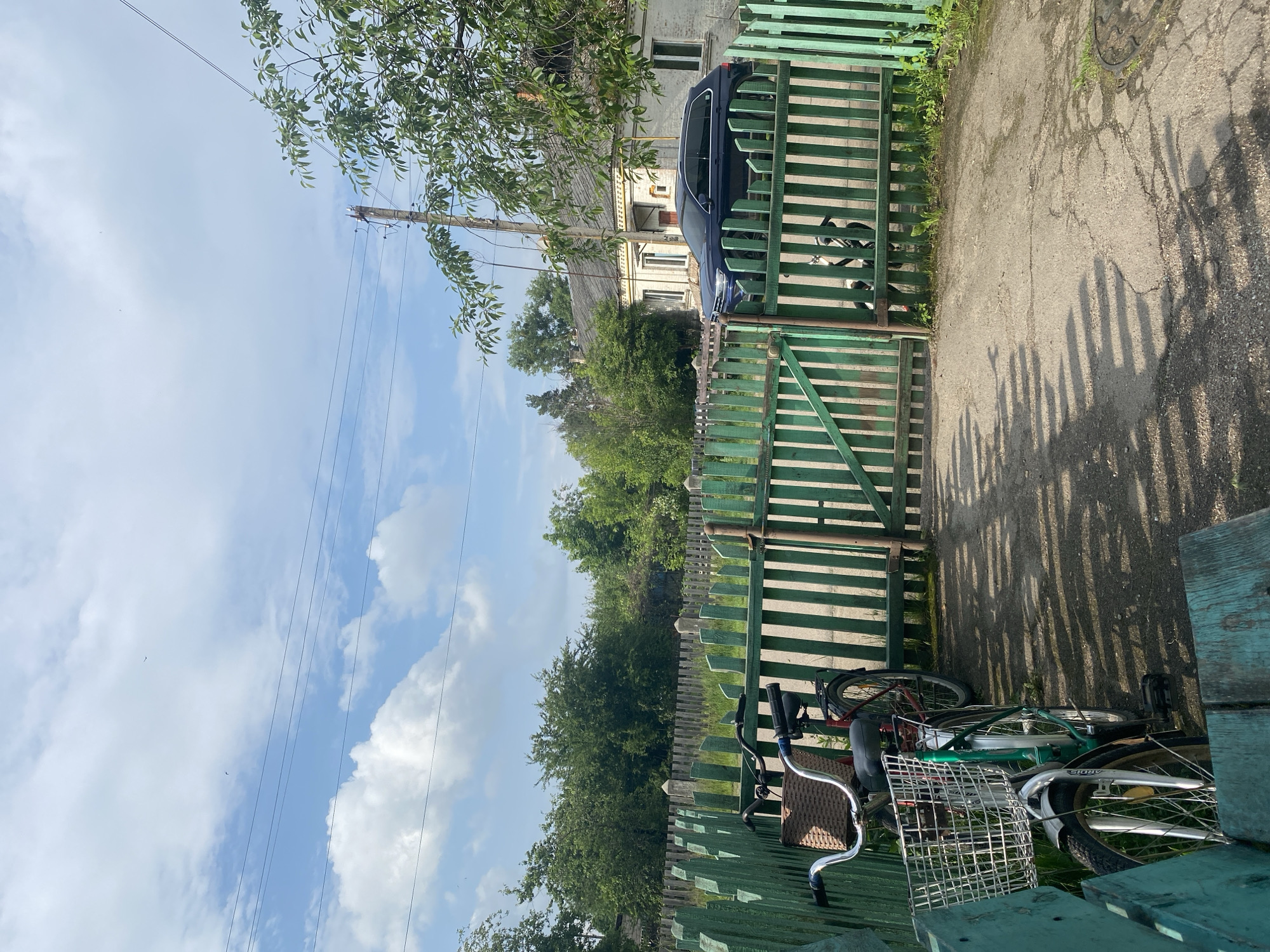 This image depicts a static outdoor scene in Ihnatpil, Ukraine, characterized by a semi-rural or village environment under a partly cloudy sky.
In the foreground, a weathered teal-green wooden bench or low structure is partially visible on the bottom left. Adjacent to it, two bicycles are parked upright next to a green wooden picket fence. The closer bicycle features a green frame, silver fenders, chrome handlebars, and a large, square silver wire basket mounted on the front. The further bicycle, with a red frame, has a brown wicker basket attached to its handlebars. Both bicycles cast long shadows extending towards the lower left.
A prominent green wooden picket fence with a matching gate spans horizontally across the midground. The gate appears slightly ajar. The ground in front of the fence is a light grey paved surface, cracked in various places, with small patches of grass growing through. Long, distinct shadows from the fence pickets fall across this paved area, indicating sunlight from the right side of the frame. A round, dark metal manhole cover is visible embedded in the pavement in the lower right corner.
Behind the green fence, an older, more weathered wooden fence with concrete posts delineates another boundary. Beyond this, dense green foliage and numerous trees form a natural backdrop, suggesting overgrown areas or a small forest.
In the background on the right, a light-colored, single-story house with a grey pitched roof is partially visible. The house has white-framed windows and decorative white trim. A dark blue SUV is parked in front of the house, largely obscured by the green fence. To the right of the house, a wooden utility pole with multiple overhead wires extends upwards. Branches with green leaves from an unseen tree overhang the upper right portion of the scene.
The upper portion of the image reveals a light blue sky with scattered white cumulus clouds. Several thin, dark lines, likely power or communication cables, traverse the sky horizontally. A few tiny, dark specks, possibly birds, are visible in the distant sky. No people are present in the scene, and no active interactions or movements are depicted.