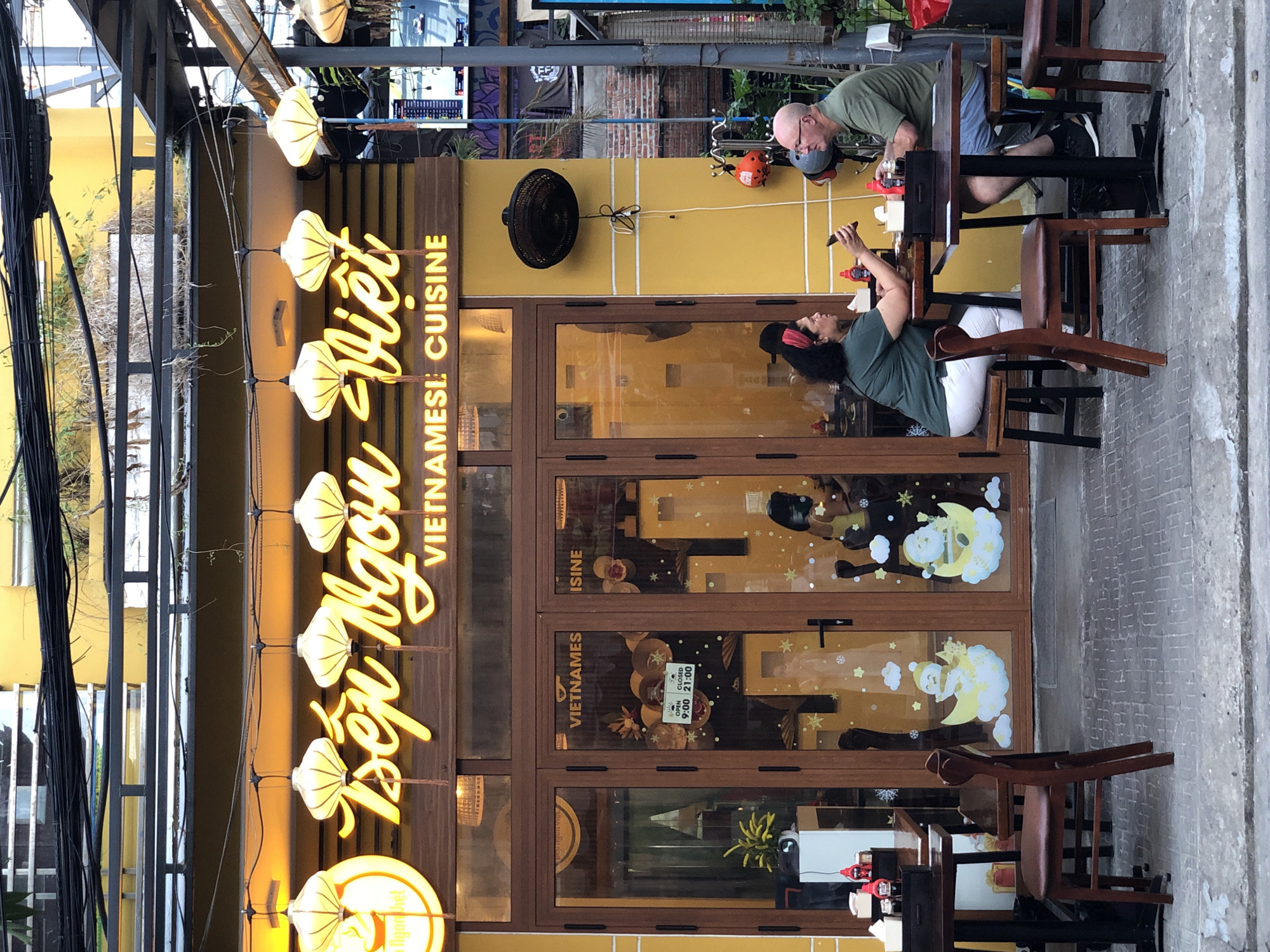 This image shows the exterior of a Vietnamese restaurant named "Bếp Ngon Việt" located in Đà Nẵng, Vietnam. The restaurant has a bright yellow facade with a large, illuminated sign featuring stylized yellow script and the text "VIETNAMESE CUISINE". Several traditional yellow lanterns are hung in front of the sign. The entrance consists of double glass doors with wooden frames.
Two patrons, a woman with dark curly hair wearing a red headband and a green shirt, and a bald man with glasses wearing a green shirt, are seated at a table outside the restaurant, facing each other. The woman is holding a smartphone. They appear to be engaged in conversation or interacting with their phones.
Other details visible include:
- Tables and chairs set up on the sidewalk for outdoor dining.
- A black oscillating fan mounted on the yellow wall.
- Decorative decals on the glass doors, including cartoon characters on moons and snowflakes.
- A sign on the door indicating "OPEN 9:00 CLOSED 21:00".
- Some greenery and overhead wires are visible.
- The ground is paved with square tiles.