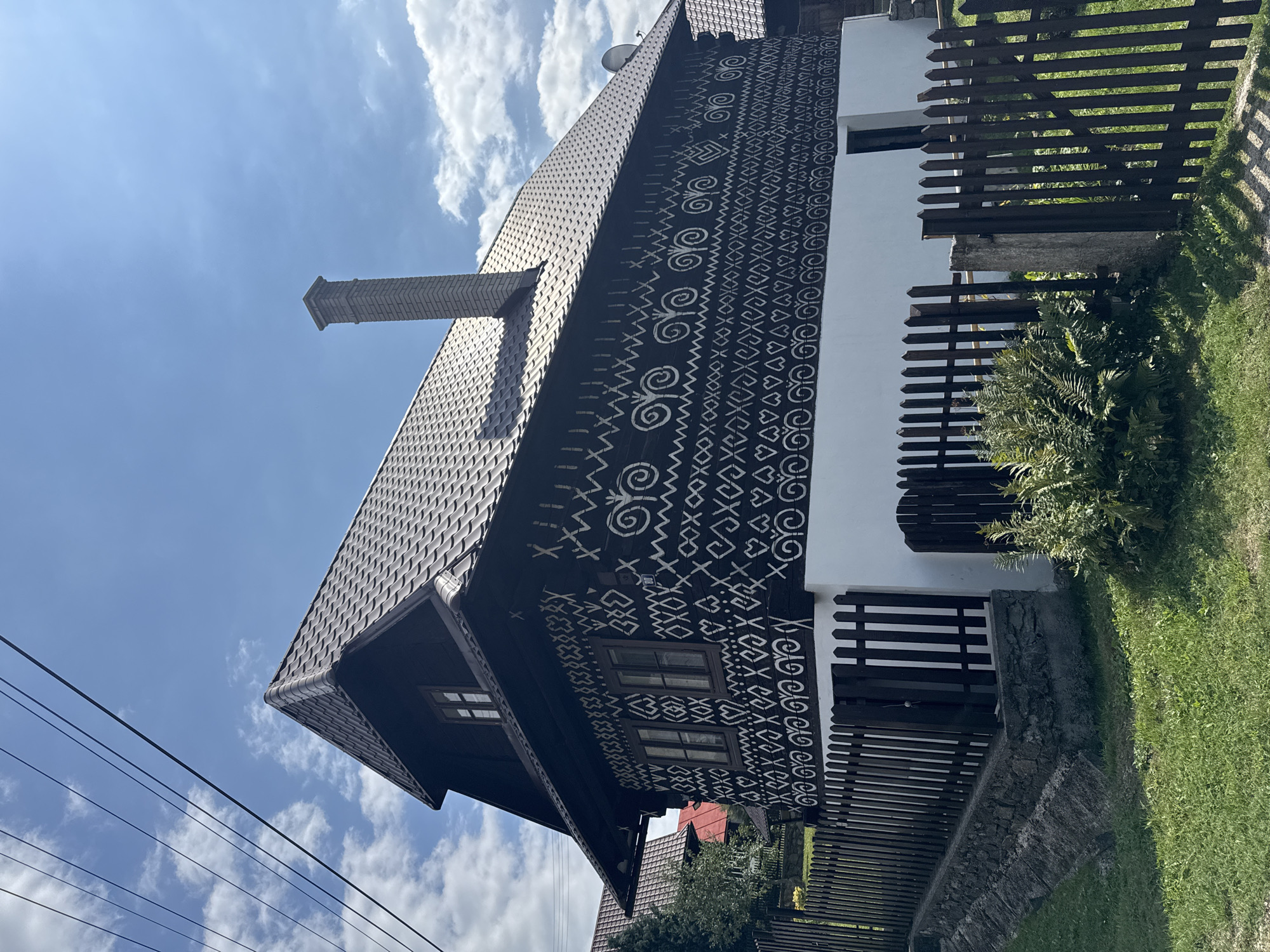 The image shows a traditional wooden house in the village of Čičmany, Slovakia. The house features dark wooden walls adorned with intricate white folk patterns. It has a dark brown tiled roof and a brick chimney. The house is partially surrounded by a wooden fence. In front of the fence, there is green grass and some green foliage. The sky is blue with some white clouds. The overall scene depicts a rural, traditional setting.