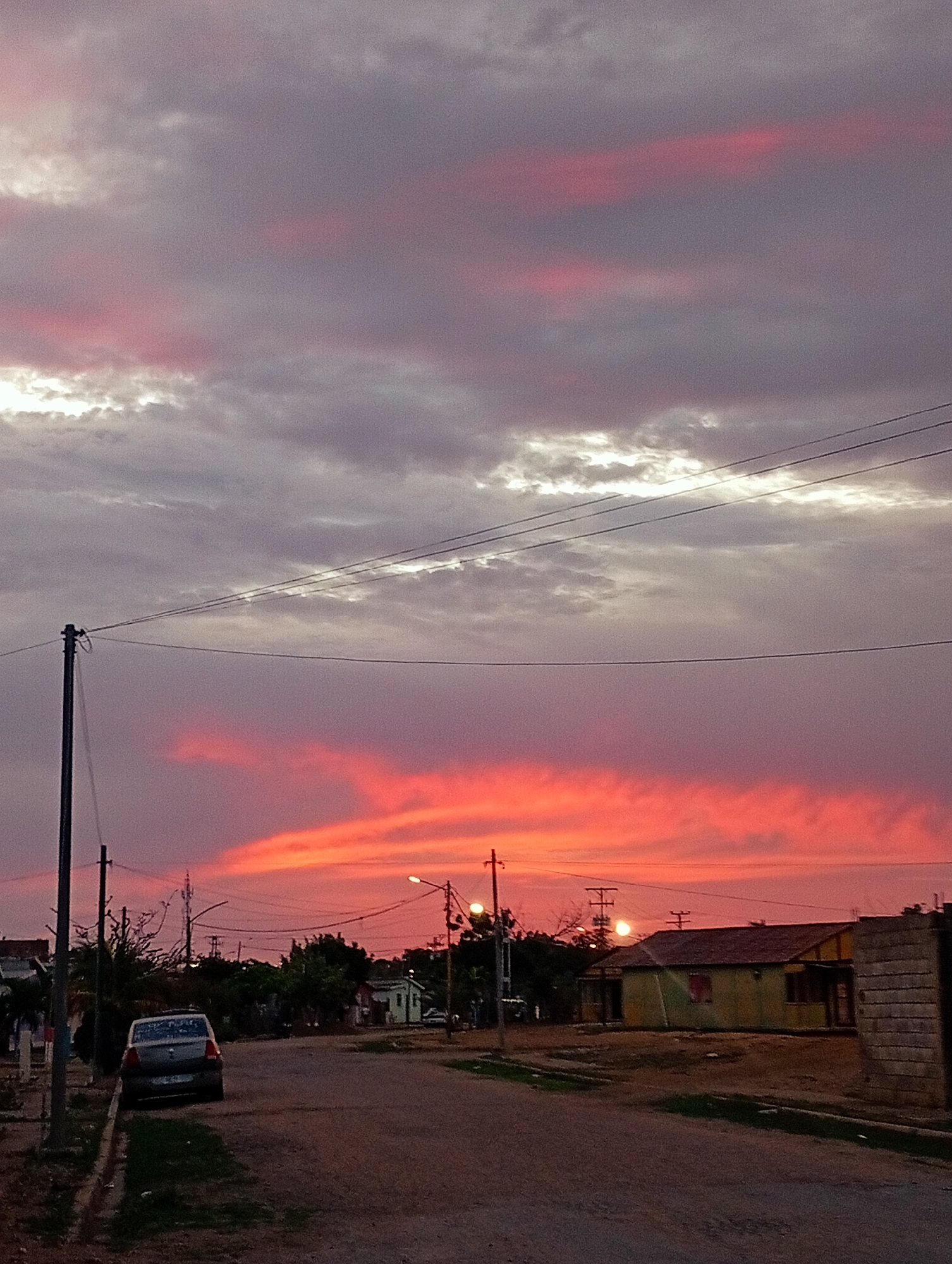 The image depicts a street scene in Juan Griego, Venezuela, during a vibrant sunset. The sky is a dramatic display of colors, transitioning from deep purples and grays in the upper portion to fiery oranges and pinks near the horizon. Clouds are scattered across the sky, catching the light of the setting sun. In the foreground, a dirt road runs diagonally from the lower right towards the center of the image. To the left of the road, a silver car is parked on the side, facing away from the camera. Adjacent to the car is a grassy area with a curb. A utility pole stands prominently on the left side of the road, with wires stretching across the frame. In the middle ground, a cluster of trees and buildings are visible. One prominent building is a long, single-story structure painted yellow with a darker roof. Streetlights are illuminated in various parts of the scene, suggesting the transition from dusk to evening. The overall environment appears to be a residential or semi-rural area, characterized by modest architecture and a quiet atmosphere. There are no people visible in the image.