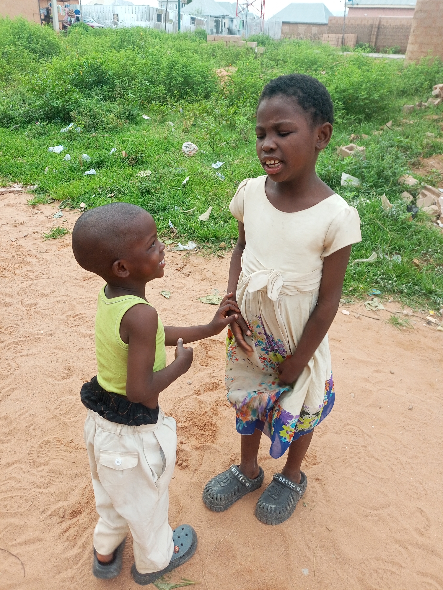 Two young children stand on a sandy ground in what appears to be a semi-urban or rural area in Tempure, Nigeria. The child on the left, a boy, is wearing a light green sleeveless shirt and light-colored pants, with a black fabric band around his waist. He has a shaved head and is looking up and to his right with his mouth open, seemingly speaking or laughing. The child on the right, a girl, is wearing a cream-colored dress with puffed sleeves and a sash tied at the waist. Her dress has a floral pattern at the hem. She is looking downwards and to her left with her eyes closed and mouth open, appearing to be speaking or reacting to something. Her dark hair is short. Both children are wearing dark-colored clogs on their feet. The environment behind them consists of overgrown green vegetation, some scattered litter, and in the distance, buildings and what might be construction materials. The ground is predominantly sandy with some footprints visible.