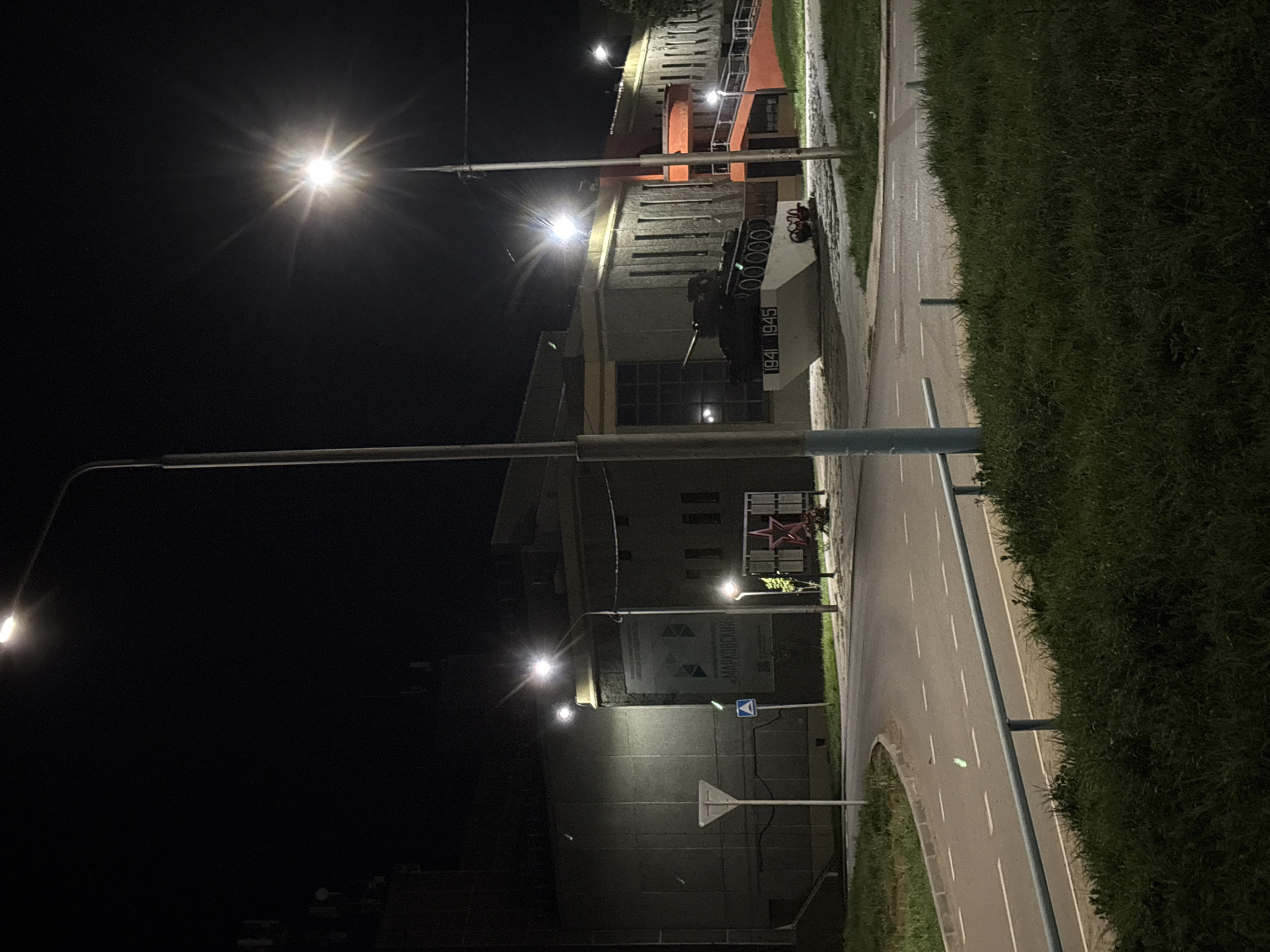 The image shows a street scene at night in Марковский, Russia. Streetlights illuminate a road with white lane markings. To the left of the road, there's a building with signage in Russian. A military tank monument stands on a pedestal near the building, bearing the dates "1941-1945." Grass and vegetation line the right side of the road. The sky is dark and featureless.