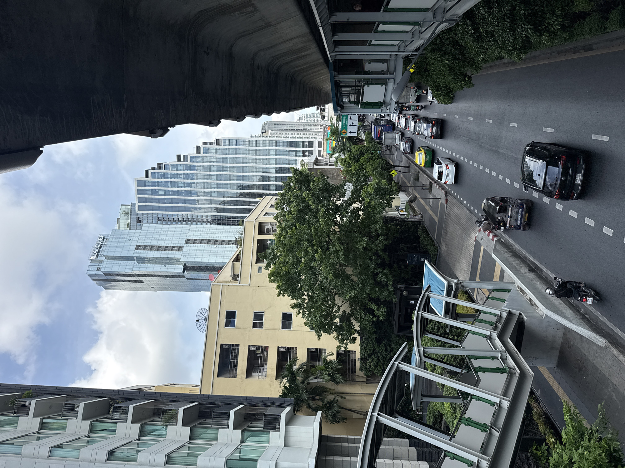 The image depicts a street scene in Bangkok, Thailand, taken from an elevated position, possibly a pedestrian bridge. The street is lined with dense traffic composed of cars, taxis, and motorcycles. The architecture includes modern skyscrapers and older, low-rise buildings, with a pedestrian overpass extending into the street. The sky is partly cloudy and a concrete structure, potentially a section of an elevated train line, dominates the upper left portion of the frame.