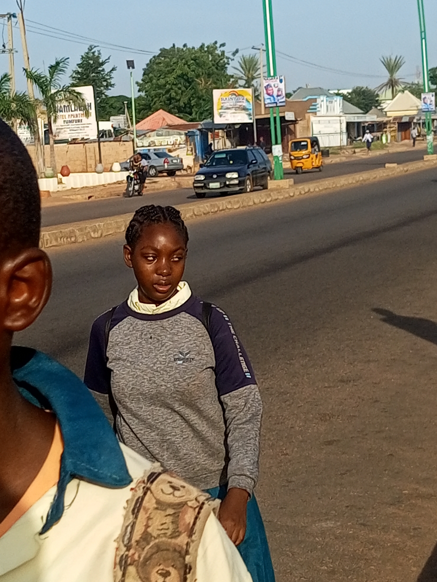 In the city of Unknown, Unknown, a young woman with braided hair walks on a paved road, her head tilted slightly as she looks to her right. She wears a grey and blue long-sleeved shirt over a light-colored garment, and a blue skirt. To her left, the shoulder and ear of another person are visible, also facing away from the camera. Behind them, a motorcycle rider is seen near a roadside structure with a large sign. Further down the road, a yellow auto-rickshaw, a dark-colored car, and several buildings are present under a clear sky. The environment appears to be a street with a sidewalk and trees lining the background.