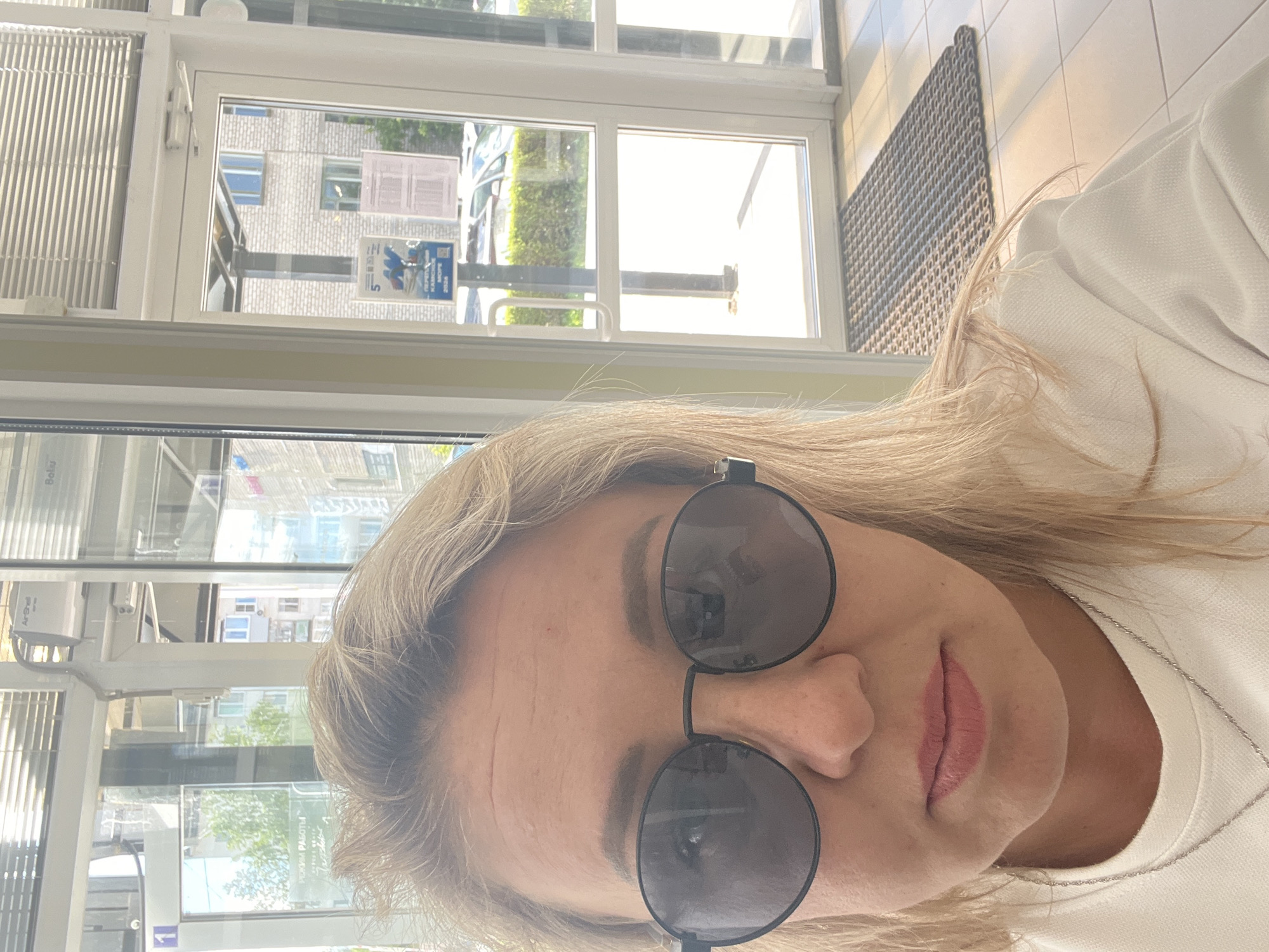 The image is a selfie of a Caucasian woman with blonde hair and sunglasses, taken indoors. She is wearing a white top and a silver necklace. The background appears to be a commercial or public building with large windows and glass doors. Through the windows, an exterior view is visible showing buildings, trees, and parked cars. The overall lighting suggests it is daytime. The text on a sign in the background is in Russian, indicating the location is likely in Russia. The presence of Russian text and the architectural style of the buildings visible outside are consistent with a location in the city of Chaikovsky, Russia.