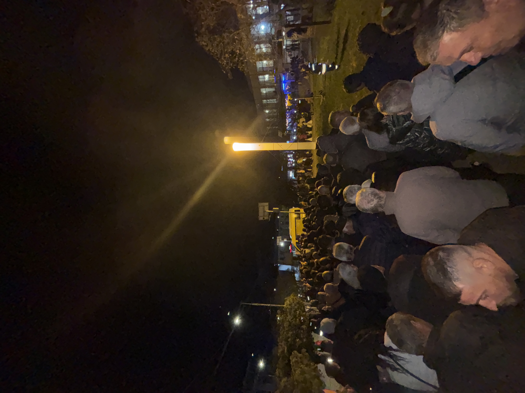 A crowd of people is gathered in Kardzhali, Bulgaria at night. A large illuminated post stands in the center of the crowd, with a building lit up in the background. Streetlights and trees line the edge of the group of people.