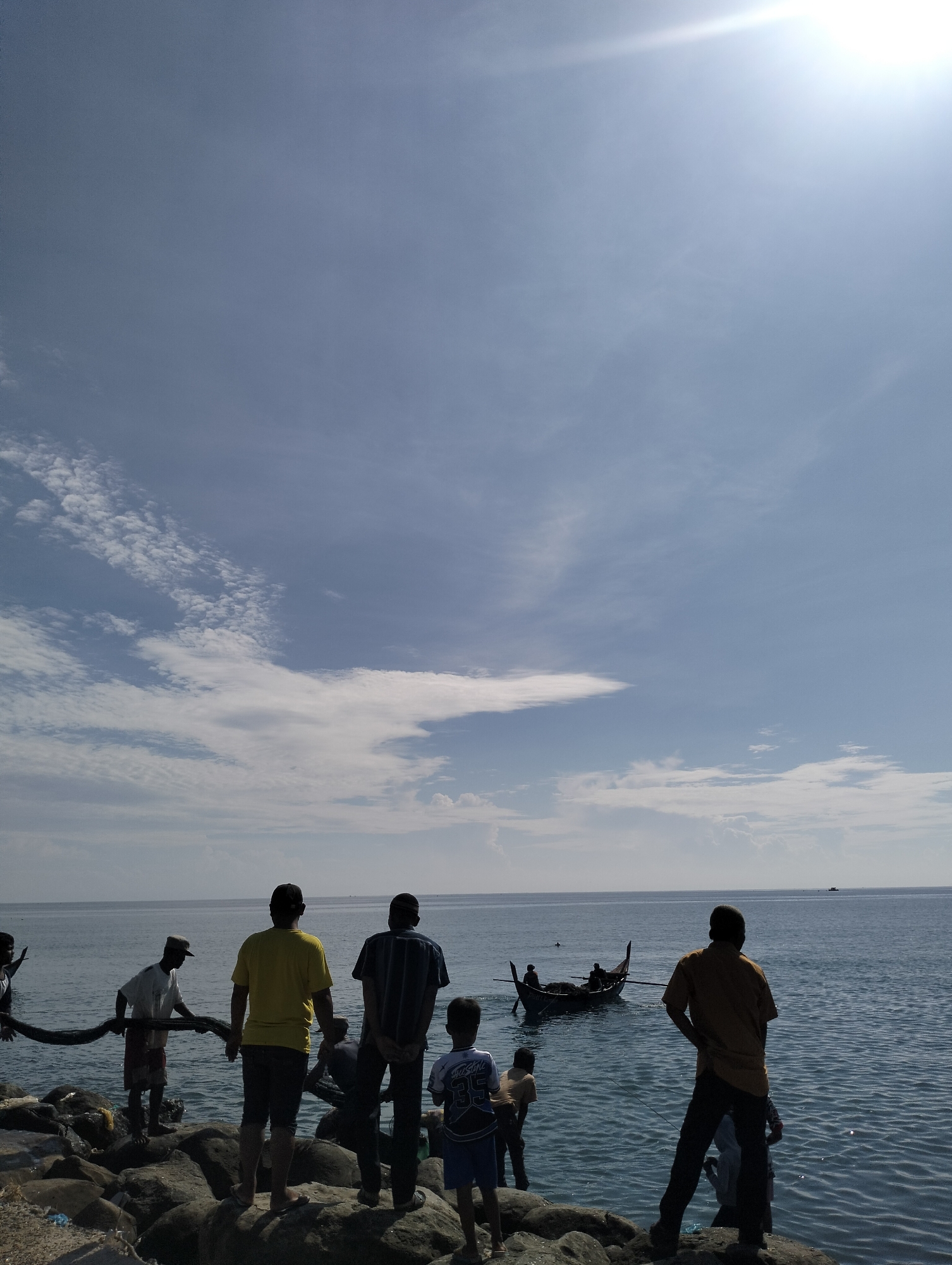 People stand on a rocky shore next to the sea. A fishing boat is visible in the water. The sky is blue with scattered clouds and a bright sun. The location is in Lhokseumawe, Indonesia.