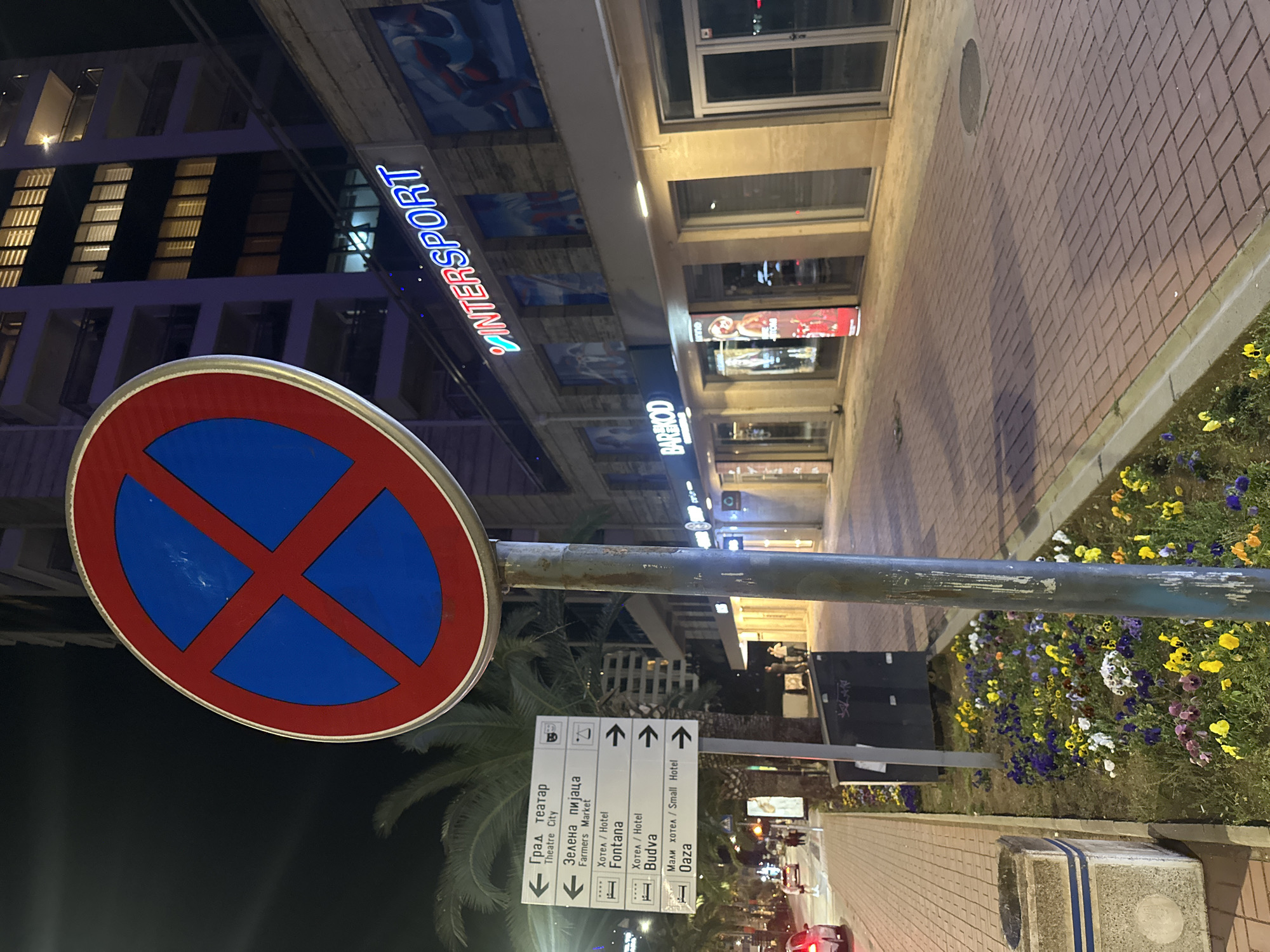 The image depicts a street scene in Budva, Montenegro at night. A no-parking sign dominates the foreground. A direction signpost indicates locations, including "Theatre City", "Farmers Market", "Fontana Hotel", "Budva Hotel", and "Oaza Small Hotel". Businesses like "INTERSPORT" and "BARIKOD" are visible. Flowers grow in a planter along the sidewalk. Vehicles are seen on the street in the distance.