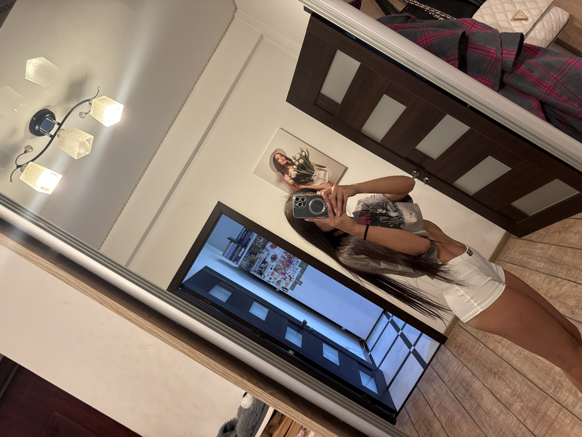 The image shows a young woman taking a selfie in a hallway. She is wearing a grey t-shirt and white shorts. The hallway has wooden floors and dark brown doors with frosted glass panels. There is a large mirror on the wall, reflecting the woman and the hallway. On the wall opposite the mirror, there is a framed picture of a woman holding a bouquet of tulips. A light fixture with four lamps is visible on the ceiling. The scene appears to be in a residential setting. There is no information to suggest this location is in Chișinău, Moldova.
