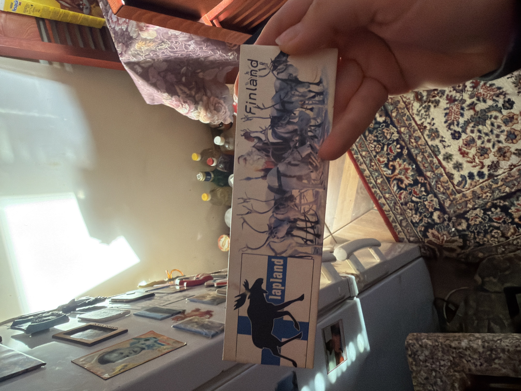A person is holding a souvenir from Finland, specifically Lapland. The souvenir features a reindeer image and the text "Finland" and "Lapland." The background shows items on a refrigerator and a rug. The location is unknown.