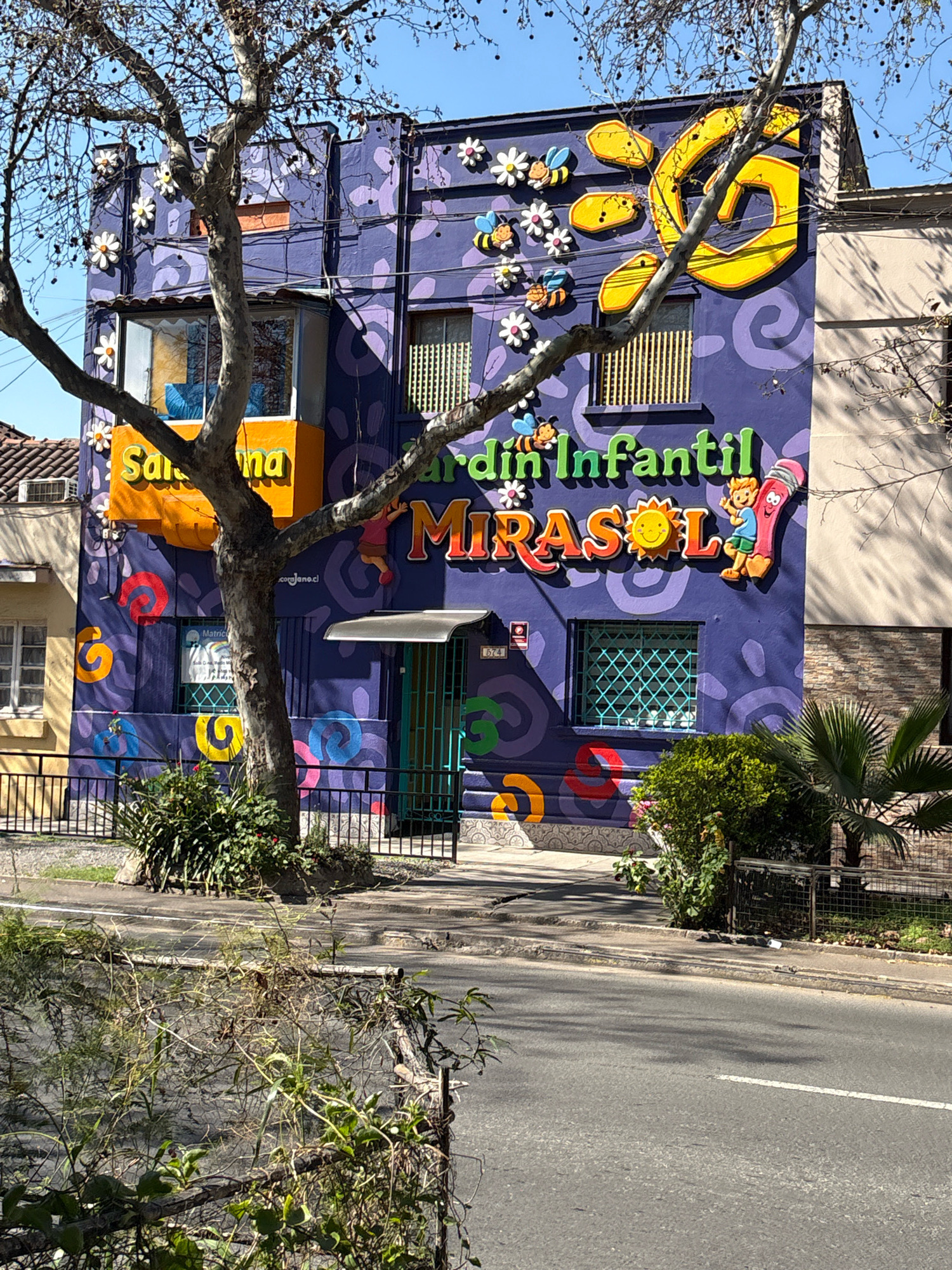 The image shows the facade of a brightly painted building with a mural. The building appears to be a kindergarten or children's center, as indicated by the text "Jardin Infantil MIRASOL" which translates to "Mirasol Kindergarten". The mural features various cartoonish elements, including flowers, bees, a sun, and a drawing of a pencil character. The building is painted in vibrant colors, predominantly purple, with yellow and orange accents. A large tree with bare branches is in the foreground, partially obscuring the building. The scene is located on a street with a sidewalk and a road. The architectural style suggests it is in a city in Chile, likely Santiago. The building has several windows, some with decorative green bars. A small sign with the number "674" is visible near the entrance. The surrounding environment includes other buildings, some with tiled roofs, and vegetation. The sky is clear and blue, indicating a sunny day.