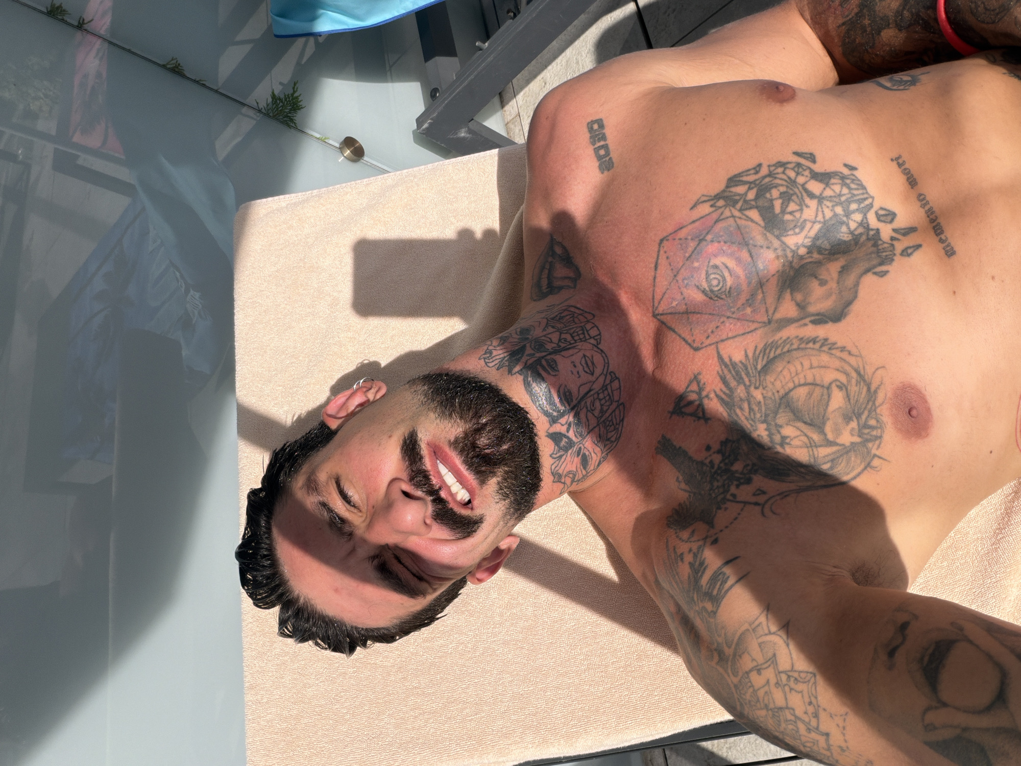 This media file depicts a man lying on a light-colored towel on what appears to be an outdoor deck or patio. The man is facing upwards, with his head tilted back. He has dark hair, a beard, and is wearing an earring in his left ear. His chest and arms are covered in tattoos, some of which are intricate designs, including a skull with geometric shapes and a dragon. There is text visible on his chest, including "2020" and "memento mori".
The scene is bathed in strong sunlight, casting distinct shadows, suggesting it is a sunny day, likely during the midday or afternoon. The background shows reflections on a glass surface, hinting at a modern architectural setting, possibly a balcony or poolside area. A blue fabric item is visible in the upper left corner. The man's expression is relaxed, with his eyes closed and a slight smile. The overall impression is one of leisure and relaxation in a warm, sunny environment.
The location context provided is Varna, Bulgaria.