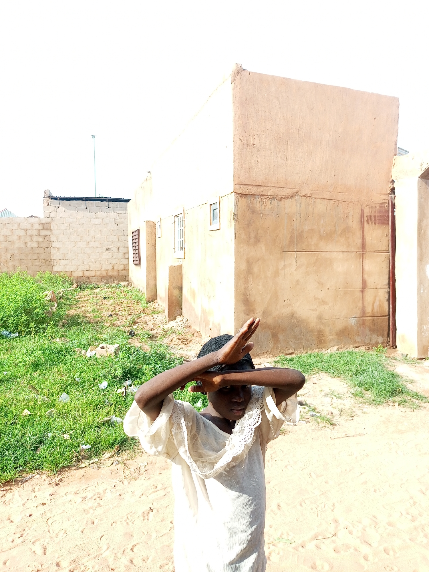 A young Black person stands in a sandy outdoor area with green grass and some debris on the left side. The person is wearing a light-colored dress and has their hands raised to block the sun from their eyes. In the background, there is a light-colored building with a rough texture and a metal gate. To the left of the building is a wall constructed of concrete blocks. The scene appears to be in a rural or semi-urban area in Tempure, Nigeria. The sky is bright and washed out, indicating a sunny day.