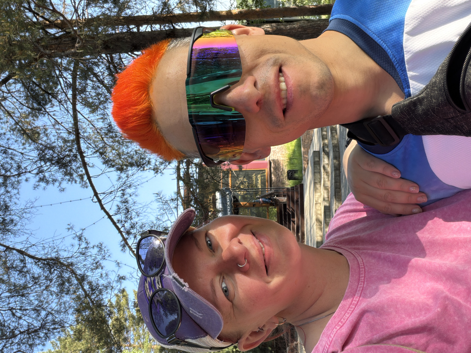 Two people are posing outdoors, likely for a selfie. The person in the upper part of the frame has bright orange hair and iridescent sunglasses. They are wearing a blue and white shirt with a black strap going across their shoulder. The person in the lower part of the frame has light skin, a nose ring, and is wearing a pink shirt with a purple baseball cap, on top of which are another pair of sunglasses. The background features trees and a blue sky. A sign can be partially seen in the background. Based on the lettering on the sign in the background, the location is most likely in the city of Chaykovsky, Russia.